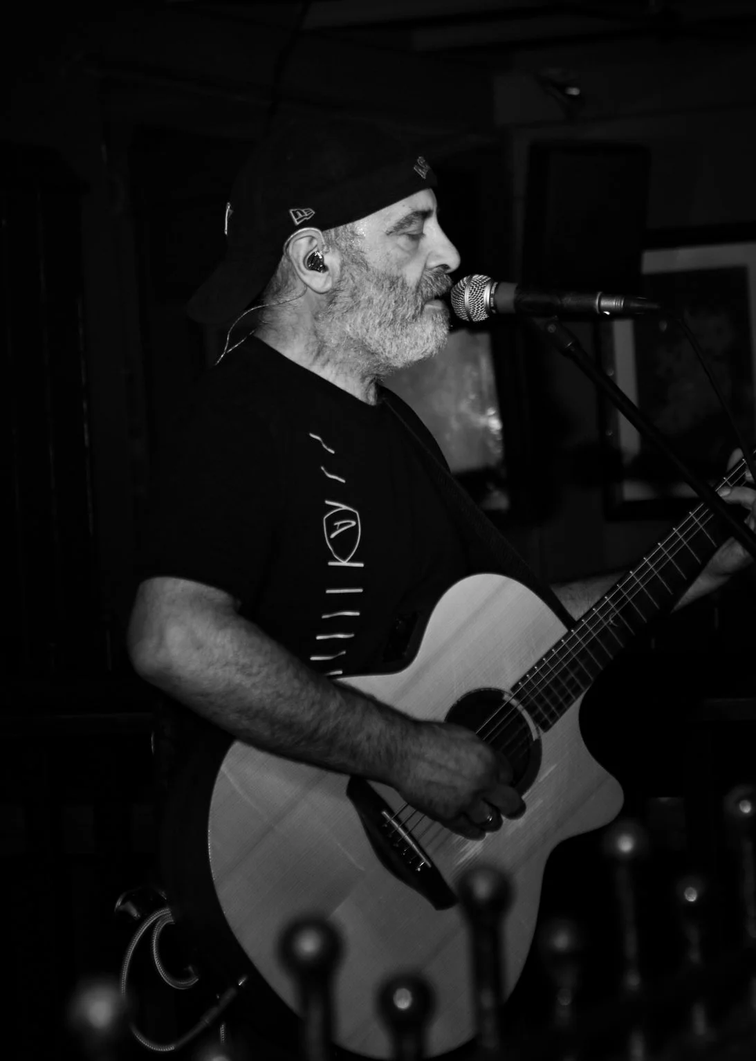 A bearded man wearing a black cap and black t-shirt playing an acoustic guitar and singing into a microphone on stage, with a dark background.