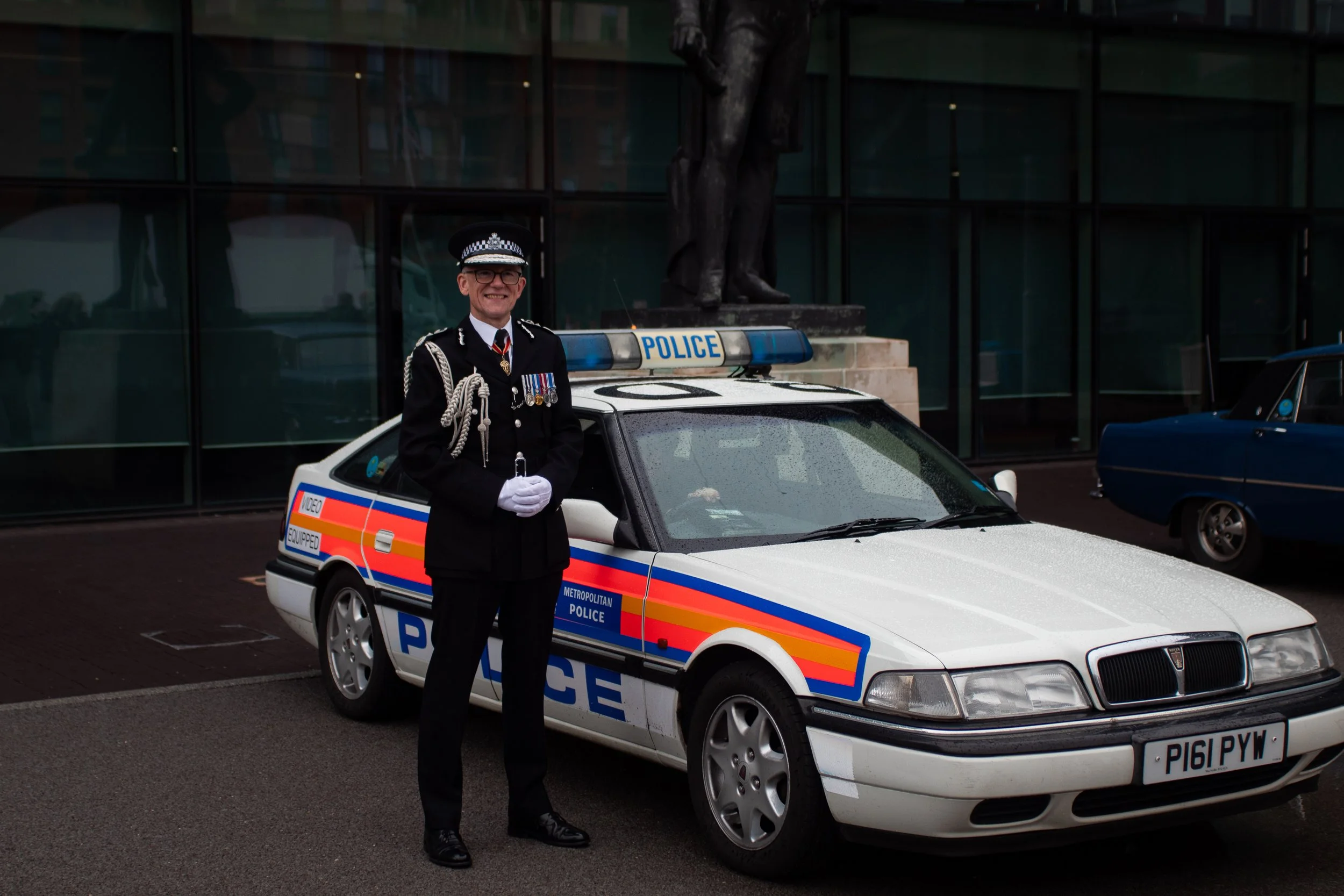 A police officer in a black uniform with medals and a peaked cap standing in front of a white police car with blue and red stripes. The car has a blue light bar and the word 'POLICE' on it. The background features a modern glass building and another 