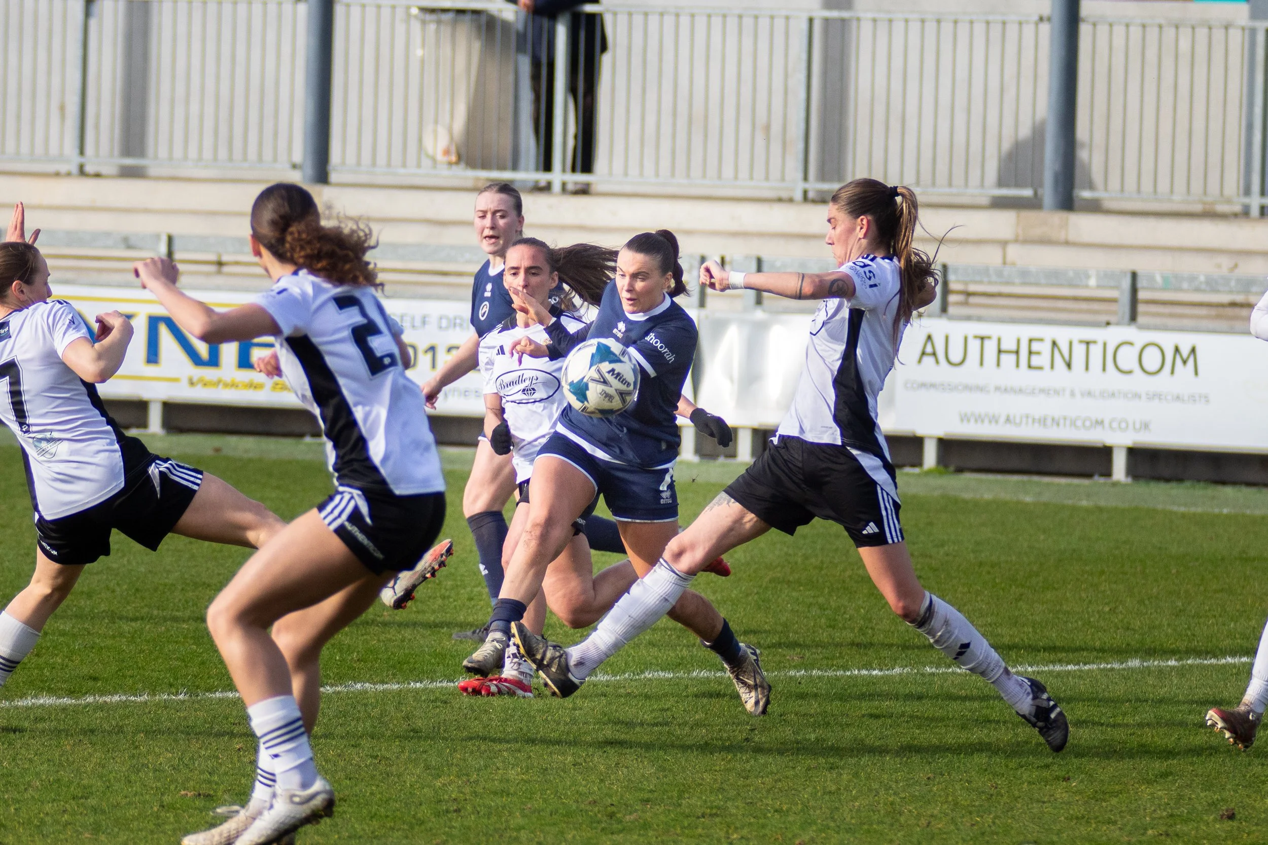 Women playing football on a field during a match, with some players tackling and others running with the ball. Millwall Lionesses