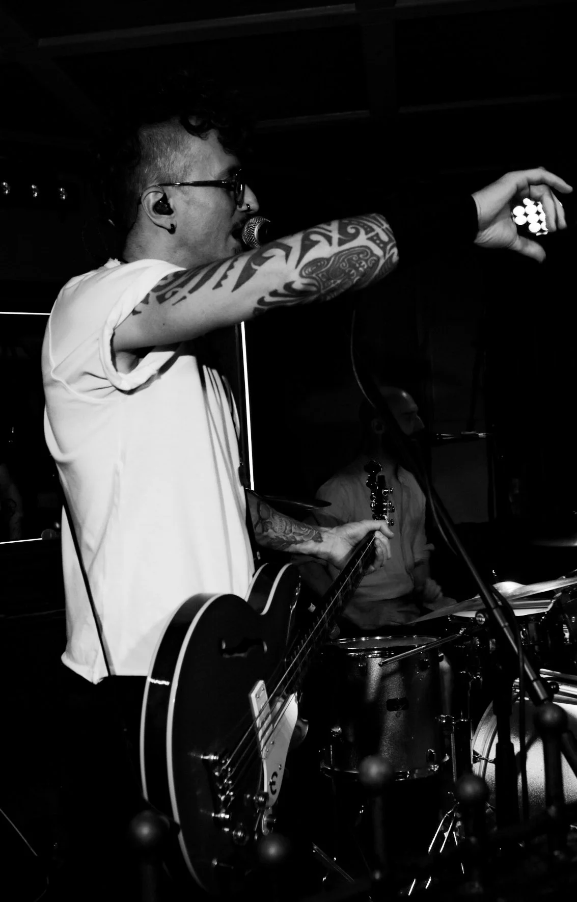 A black and white photo of a musician singing into a microphone while playing an electric guitar, with another musician playing drums in the background in a dimly lit setting.