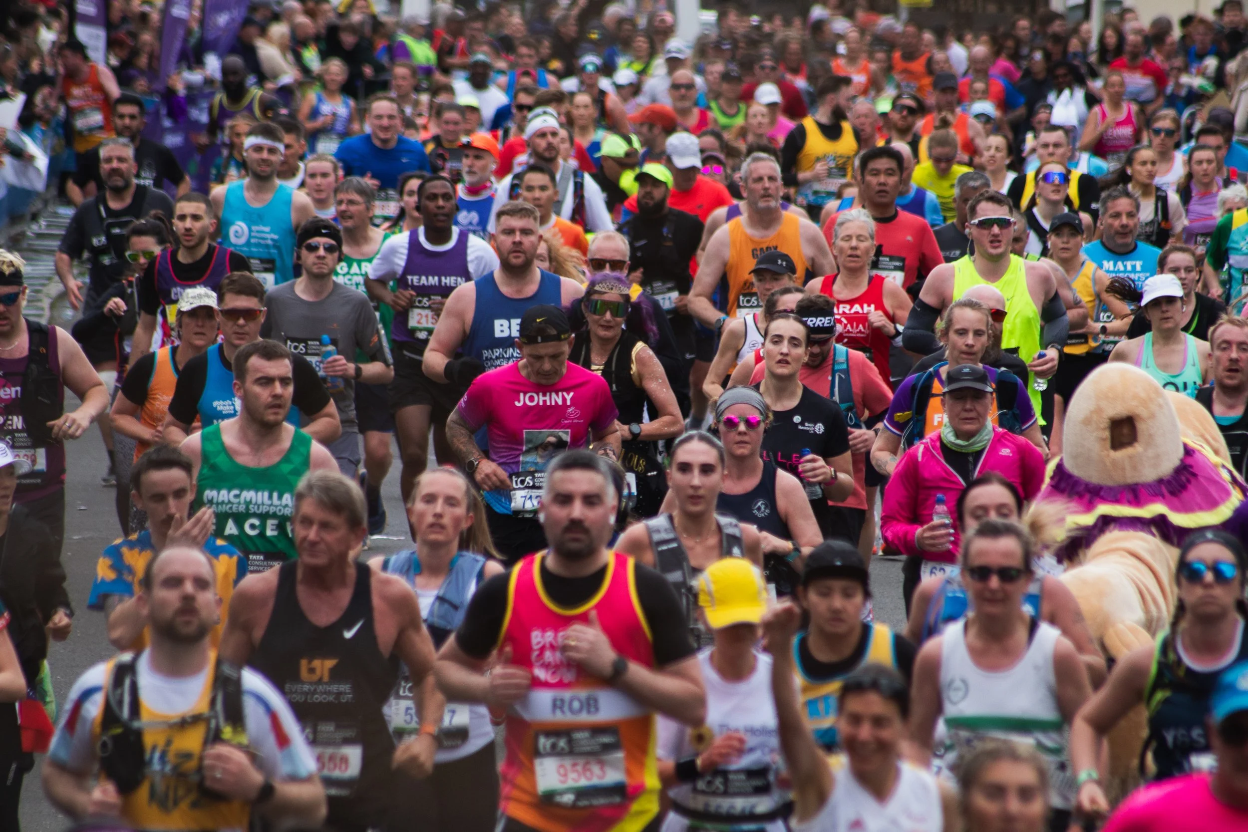 Large crowd of London marathon runners participating in a race, wearing colorful athletic clothing and accessories, moving along a street.