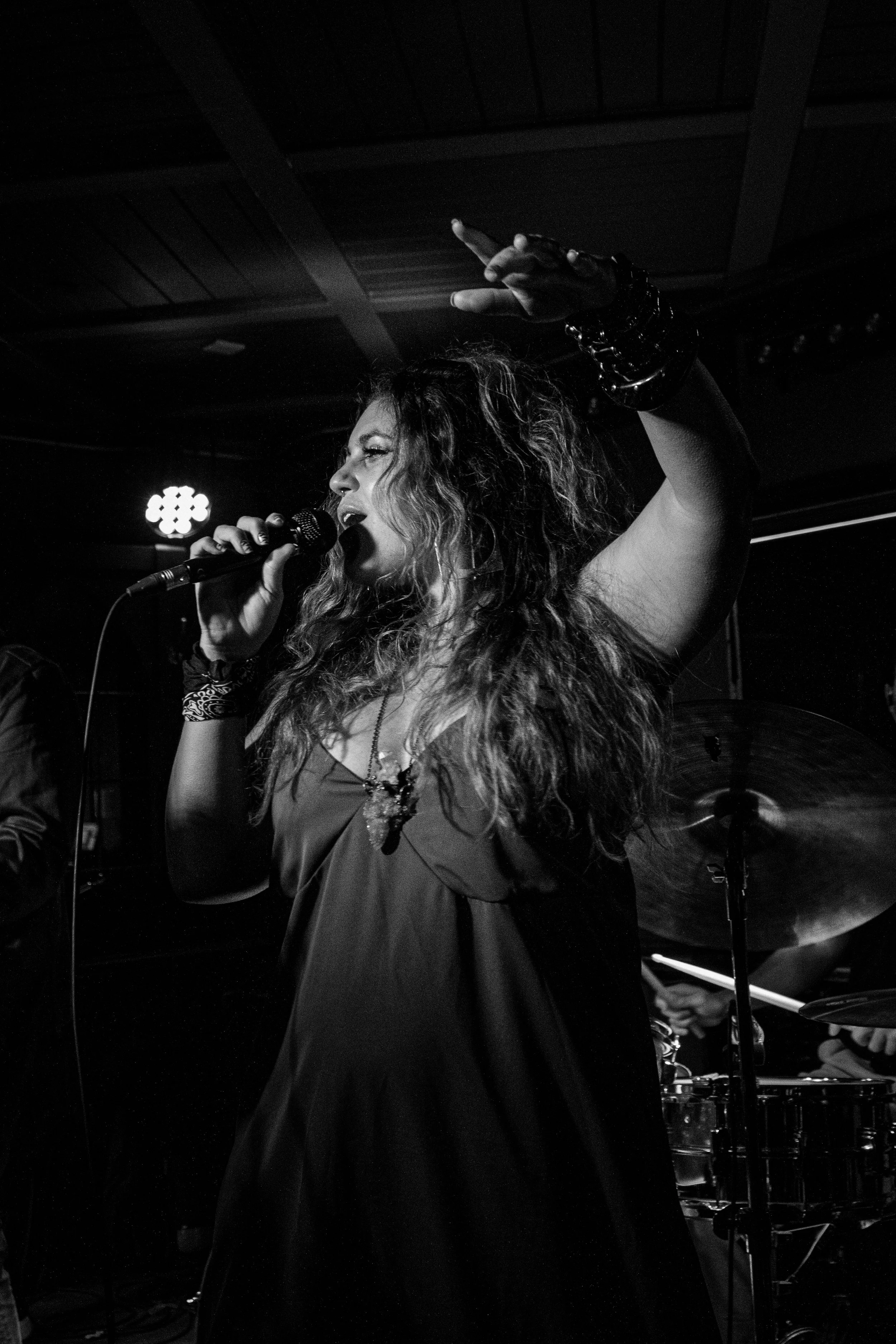 Black and white photo of a woman singing into a microphone on stage, with a drummer partially visible behind her, in a dimly lit setting.