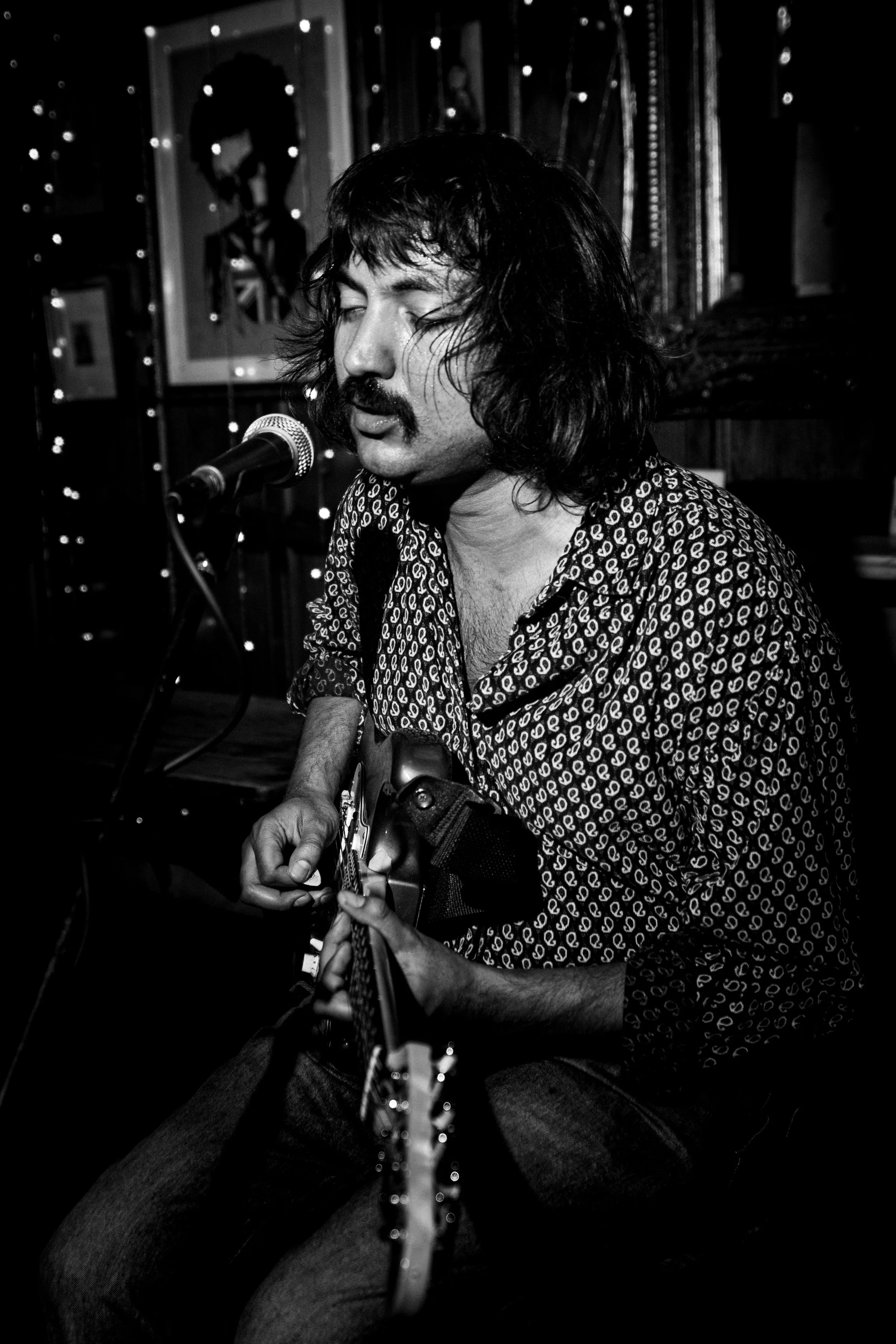 Black and white photo of a man with shoulder-length hair and a mustache, performing with a guitar in a dimly lit room, with a picture of a person's face hanging on the wall behind him. The Hawley Arms, Camden