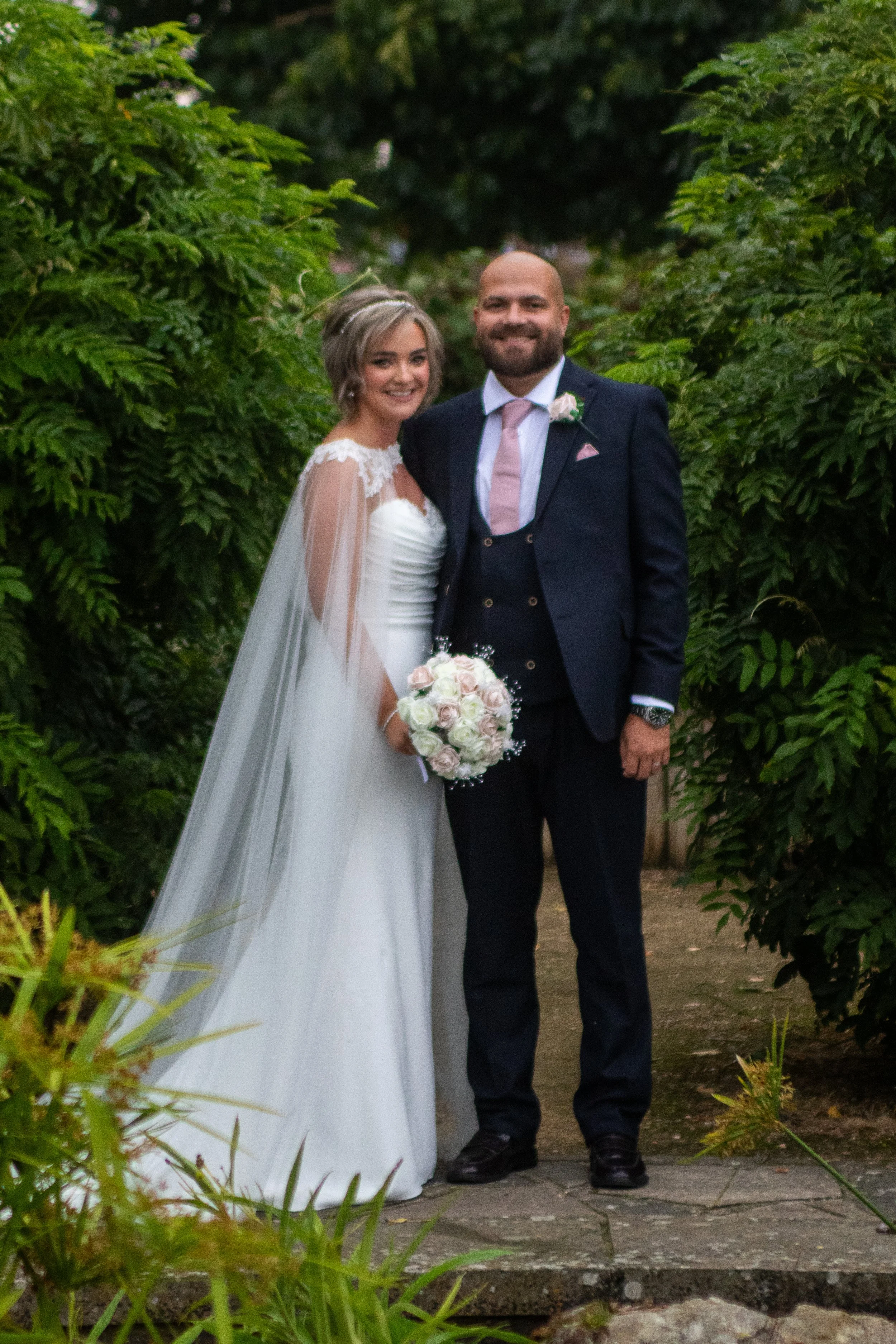 A bride and groom standing together outdoors surrounded by green foliage, smiling at the camera.