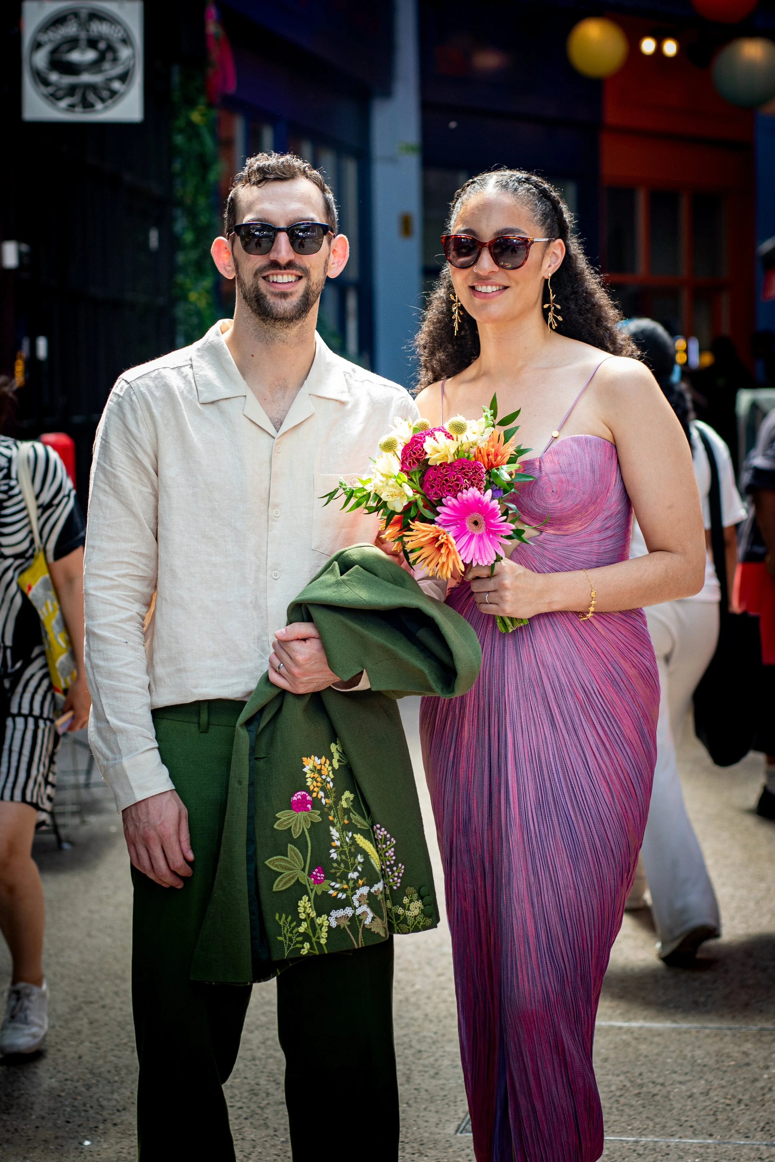 A smiling man wearing a beige shirt and sunglasses stands next to a smiling woman in a pink dress holding a colorful bouquet of flowers. They are outdoors at a festive event with colorful lanterns overhead.
