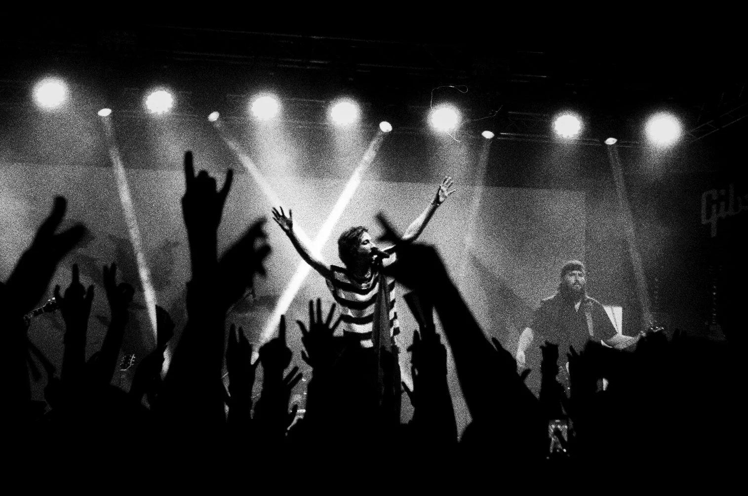 A black and white photo of a live concert with a singer on stage raising arms, surrounded by raised hands of the audience, and another musician playing guitar. At Bludfest. RUWorried