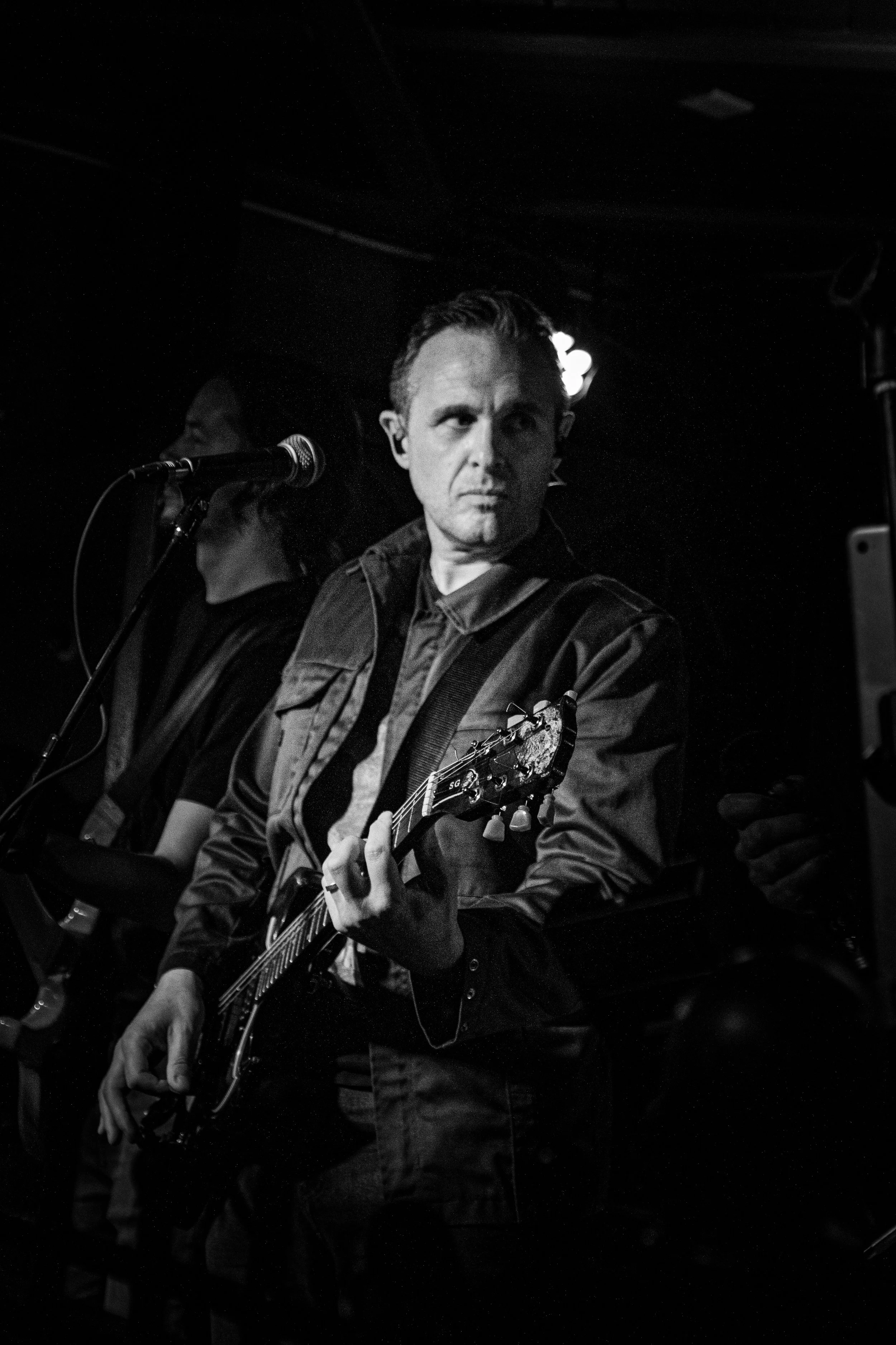 Black and white photo of a man playing an electric guitar on stage with another person in the background, singing into a microphone.