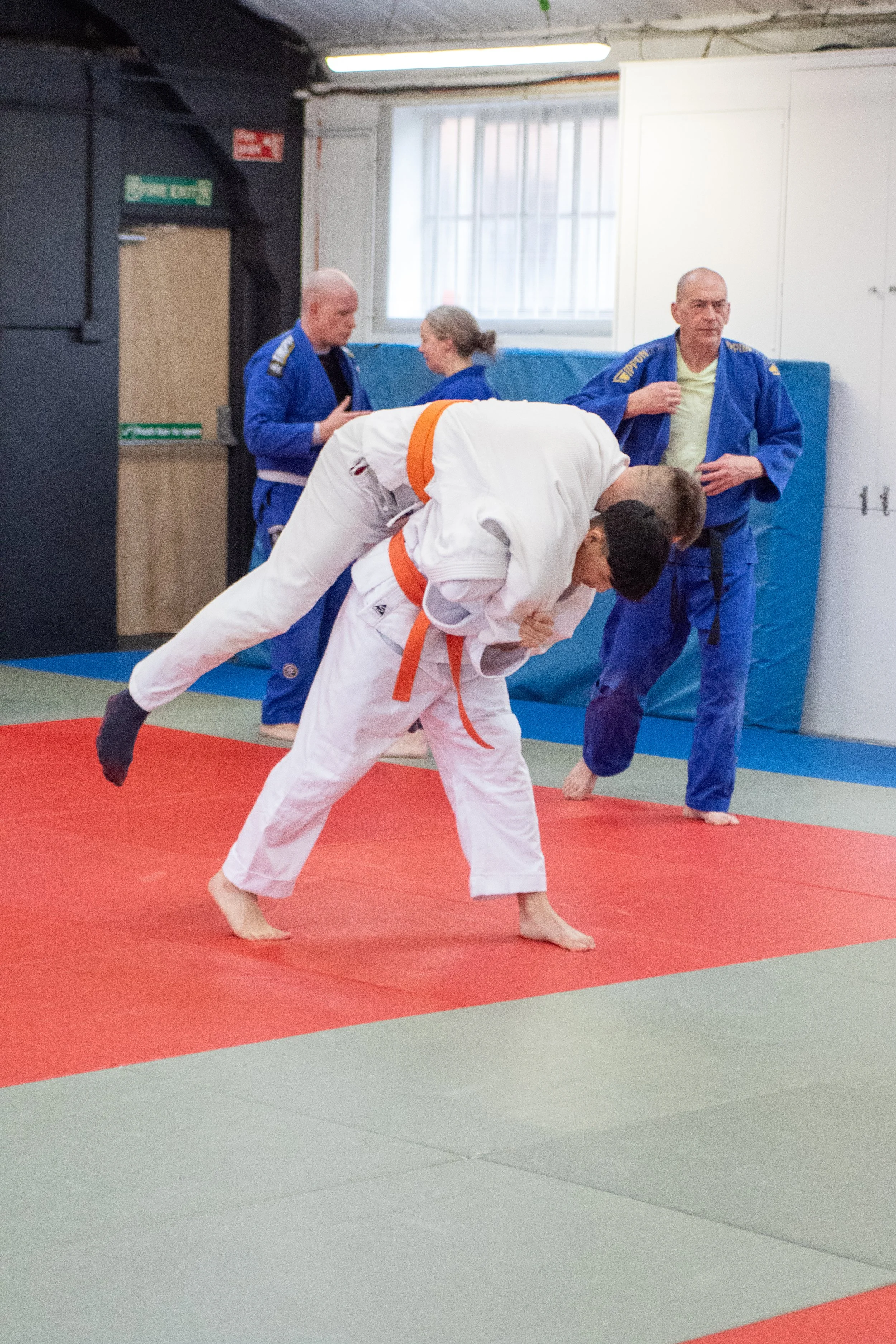 Two judo practitioners in white and blue gis practicing on a mat, with two observers in blue gis in the background. Relentless Judo, British Judo
