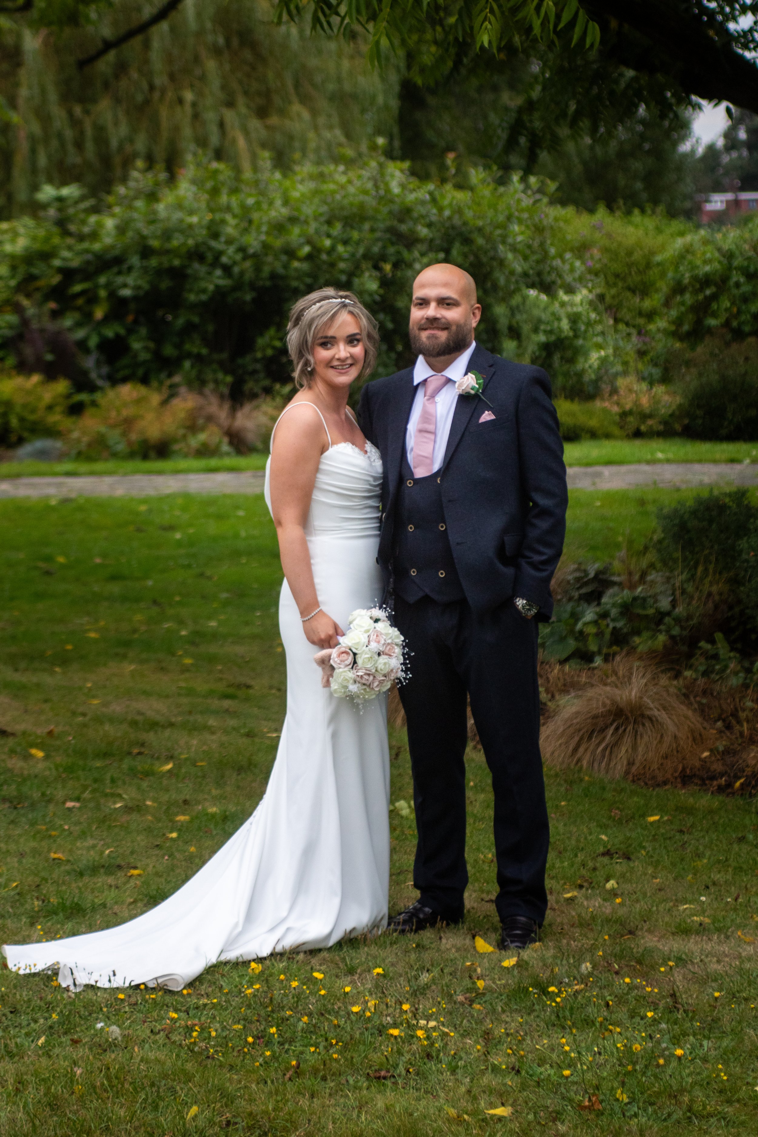 A bride and groom standing together outdoors in Danson garden, smiling for a photo. The bride is in a white wedding dress holding a bouquet of flowers, and the groom is in a dark suit with a pink tie and boutonniere. 