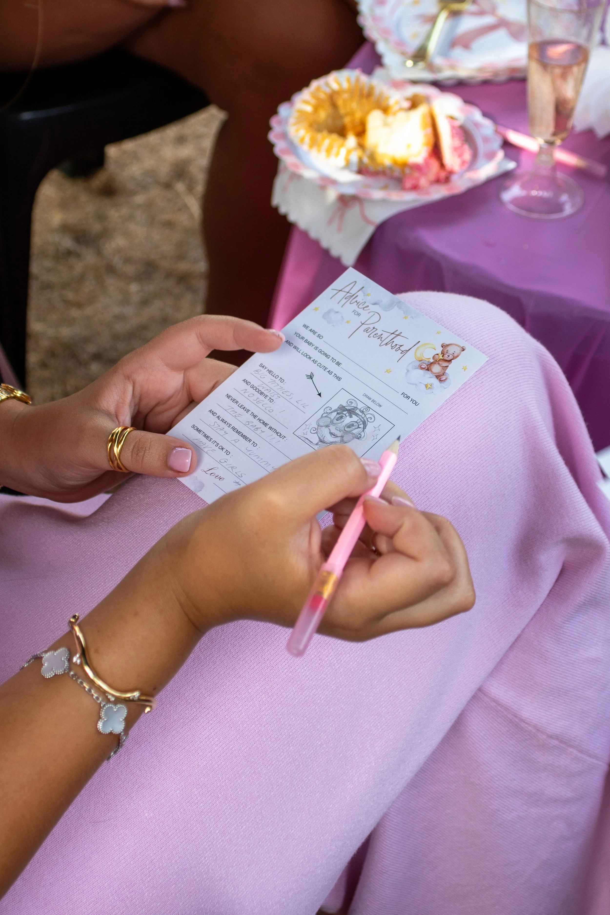 A person wearing pink pants holding an advice for parenthood card with a pink pen. In the background, there is a plate with cake and a glass of water on a pink tablecloth.