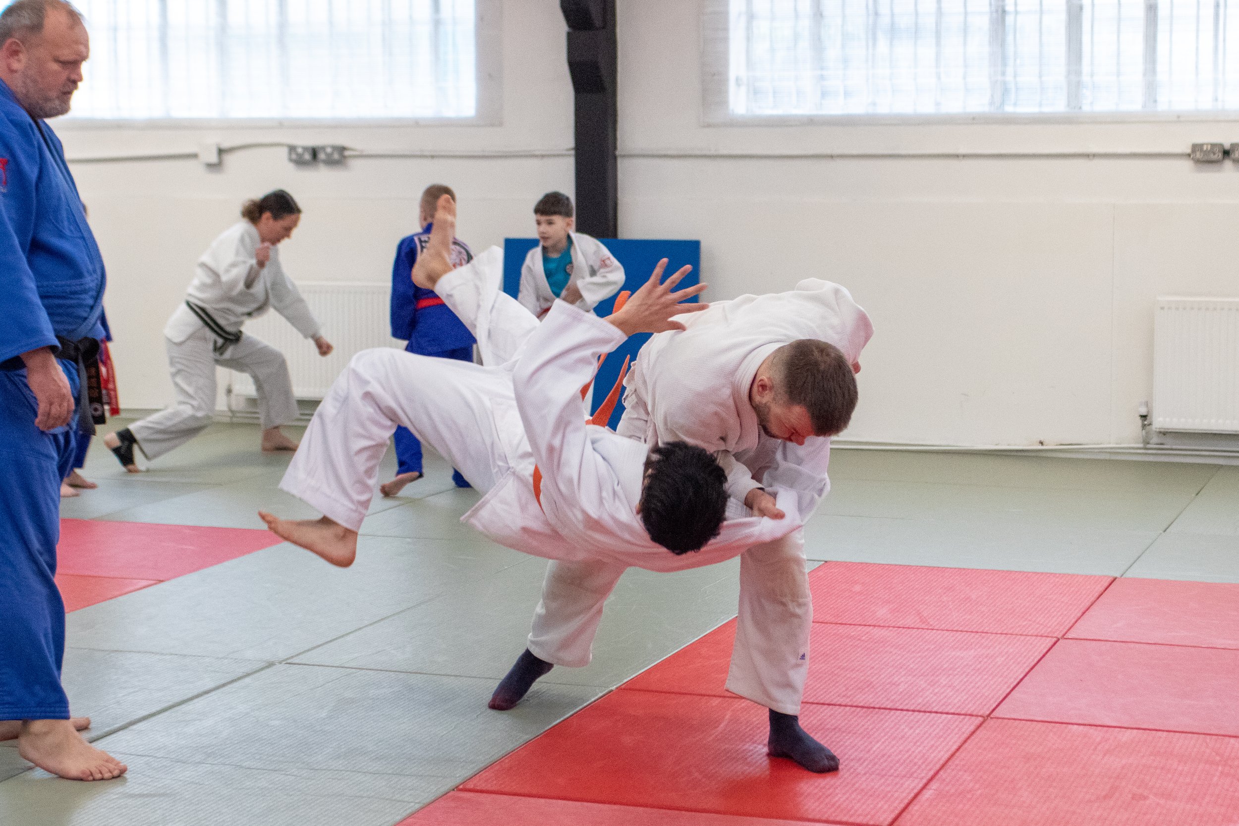 People practicing judo on mats in a dojo, with one person in white uniform executing a move while others observe or prepare to practice. Relentless Judo, British Judo