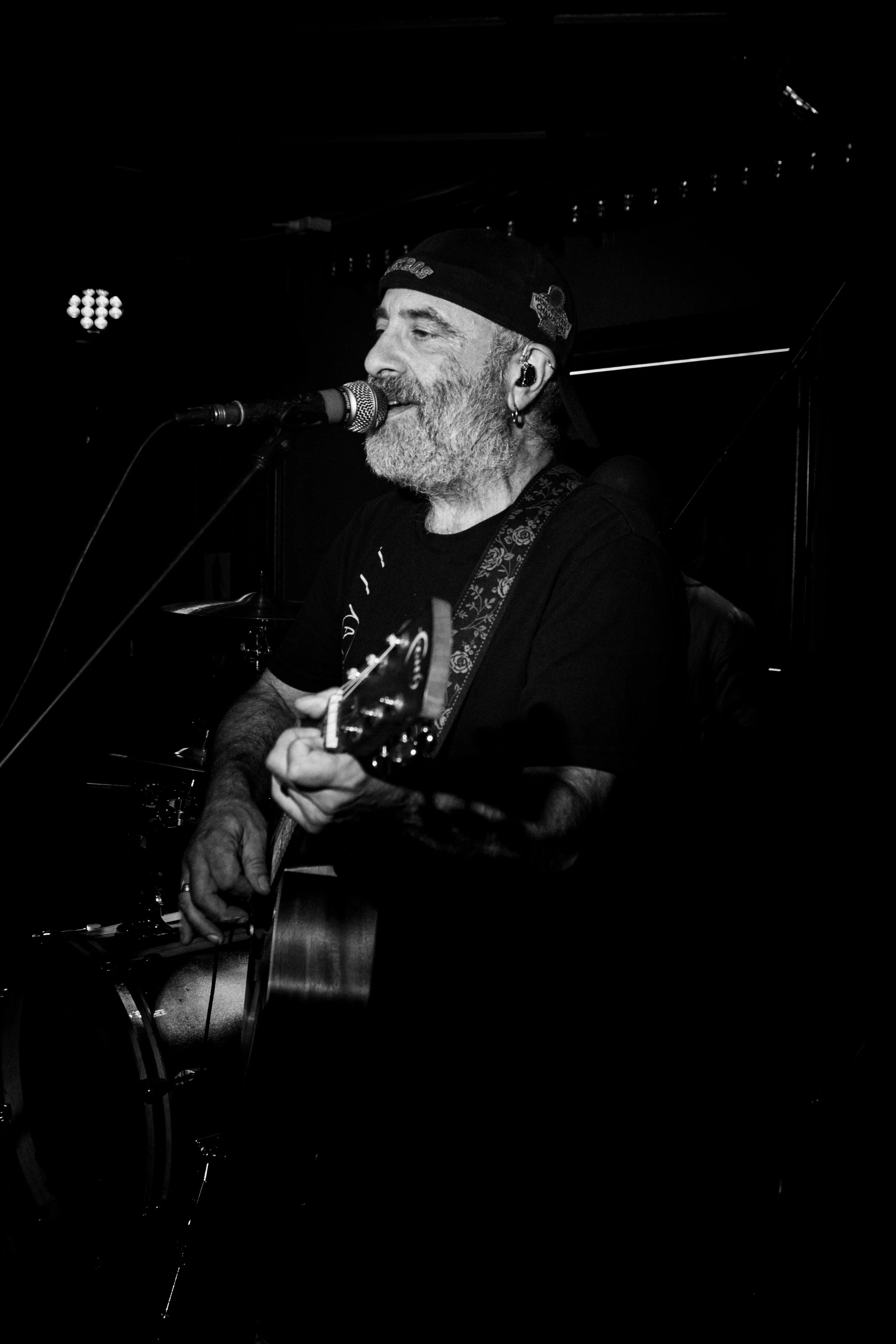 A middle-aged man with a beard, wearing a cap, playing an acoustic guitar and singing into a microphone during a live performance in a dark indoor setting.
