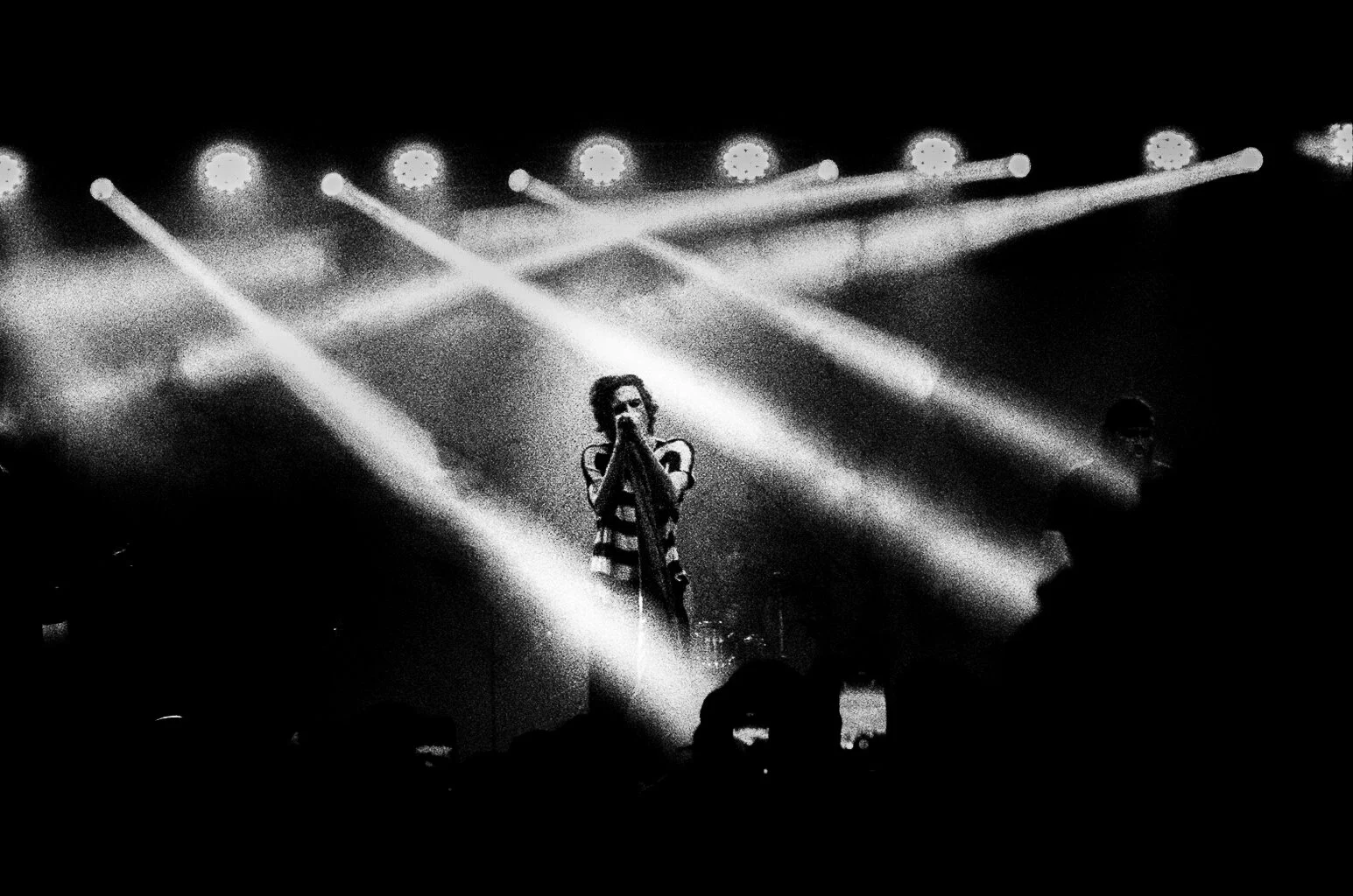 Black and white photo of a performer on stage with multiple bright lights shining down, wearing a striped outfit and holding a microphone. At Bludfest. RUWorried