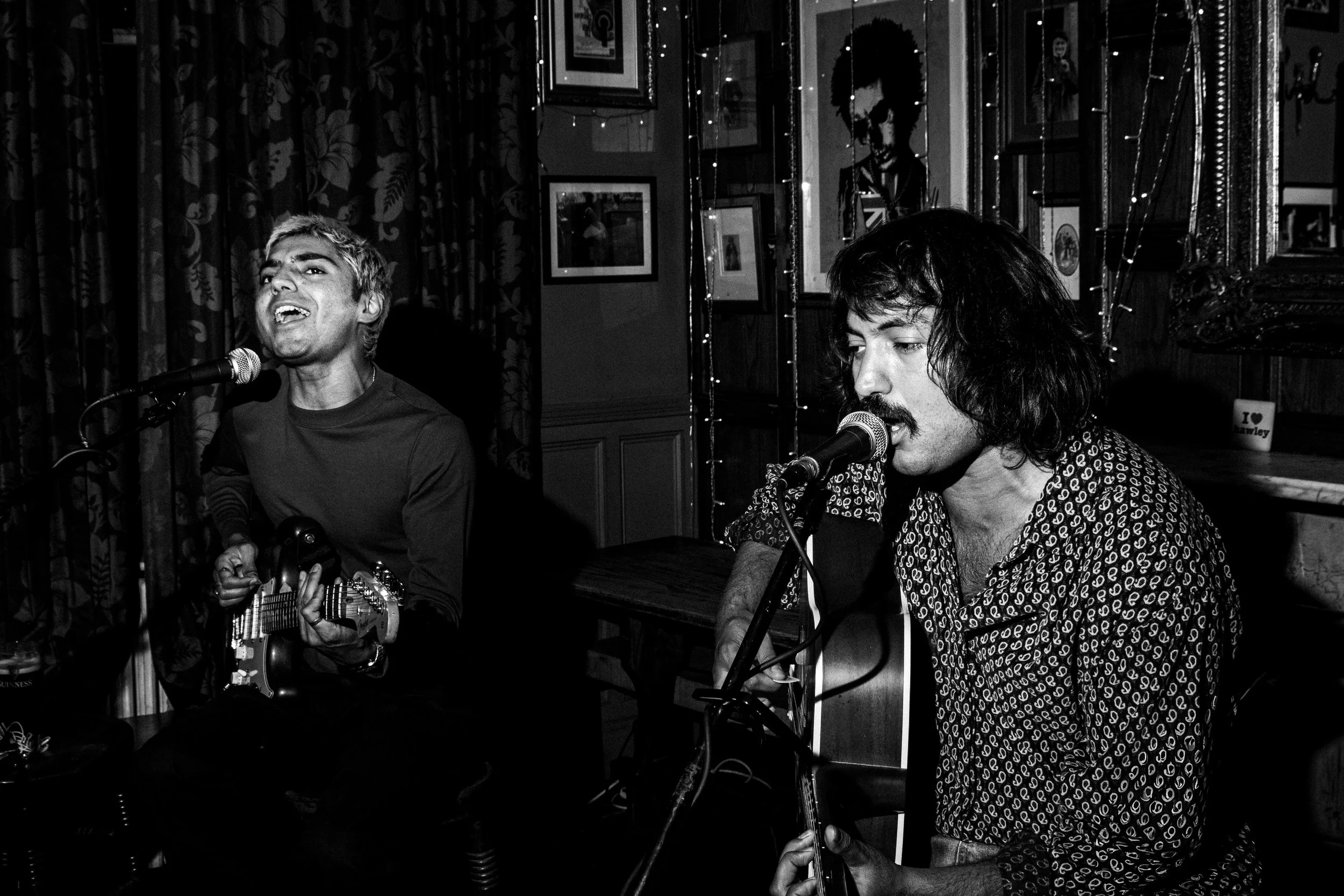 Two male musicians singing into microphones, one playing a guitar, performing in a cozy, decorated indoor venue with framed pictures and string lights. The Hawley Arms, Camden