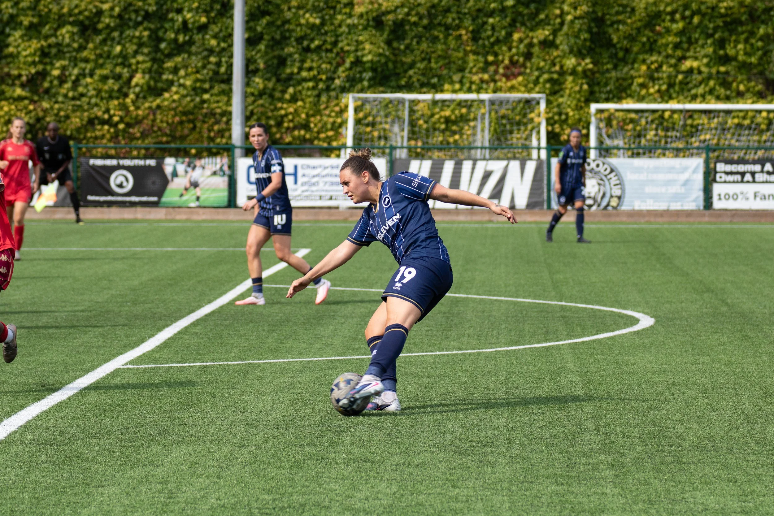 A women's football match featuring players on the field, with one player in a blue uniform kicking the ball. Millwall Lionesse Ella Ruthford and GK Barrys girlfriend