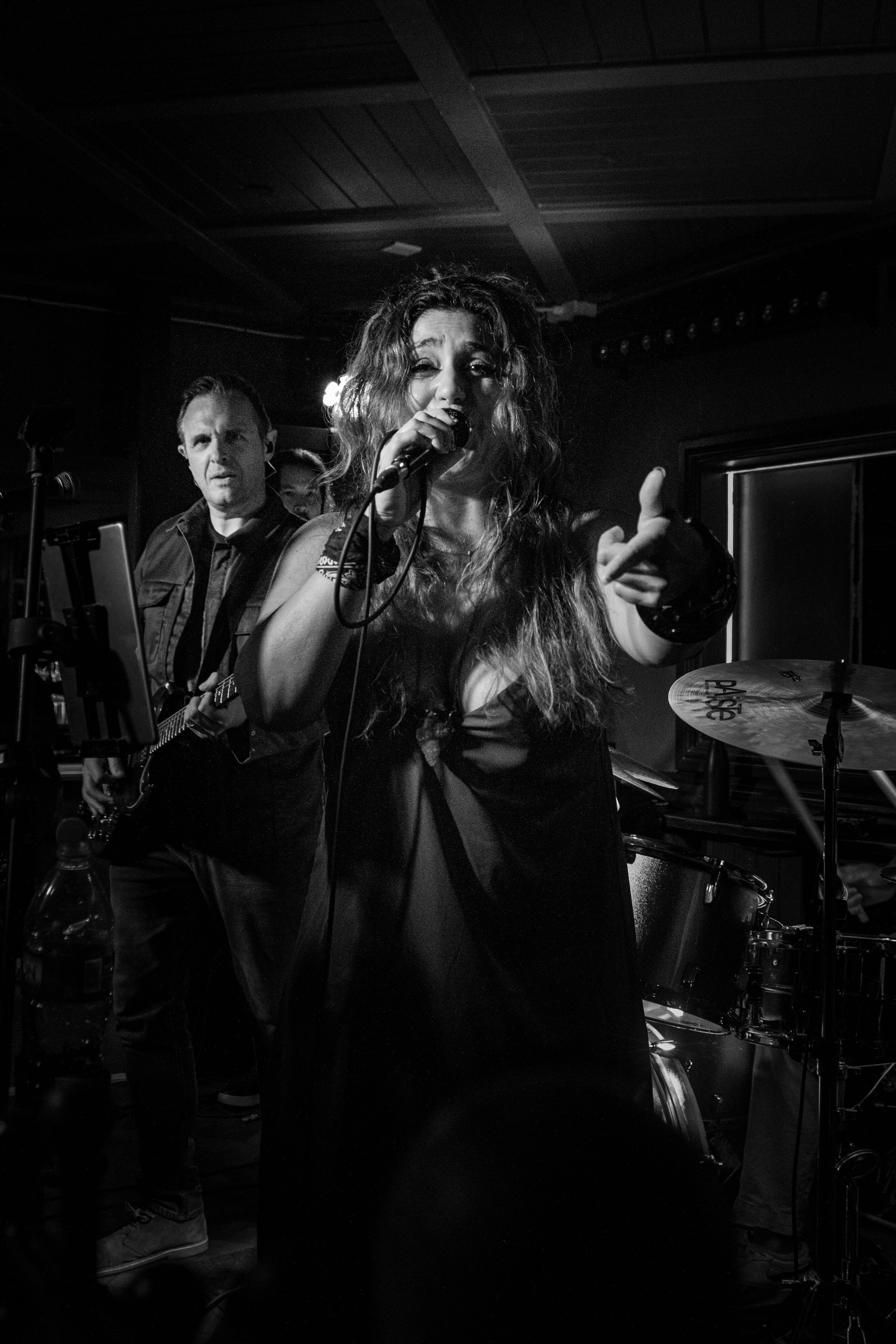 A woman singing into a microphone on stage with a drummer and a man playing guitar behind her, in a dimly lit setting.