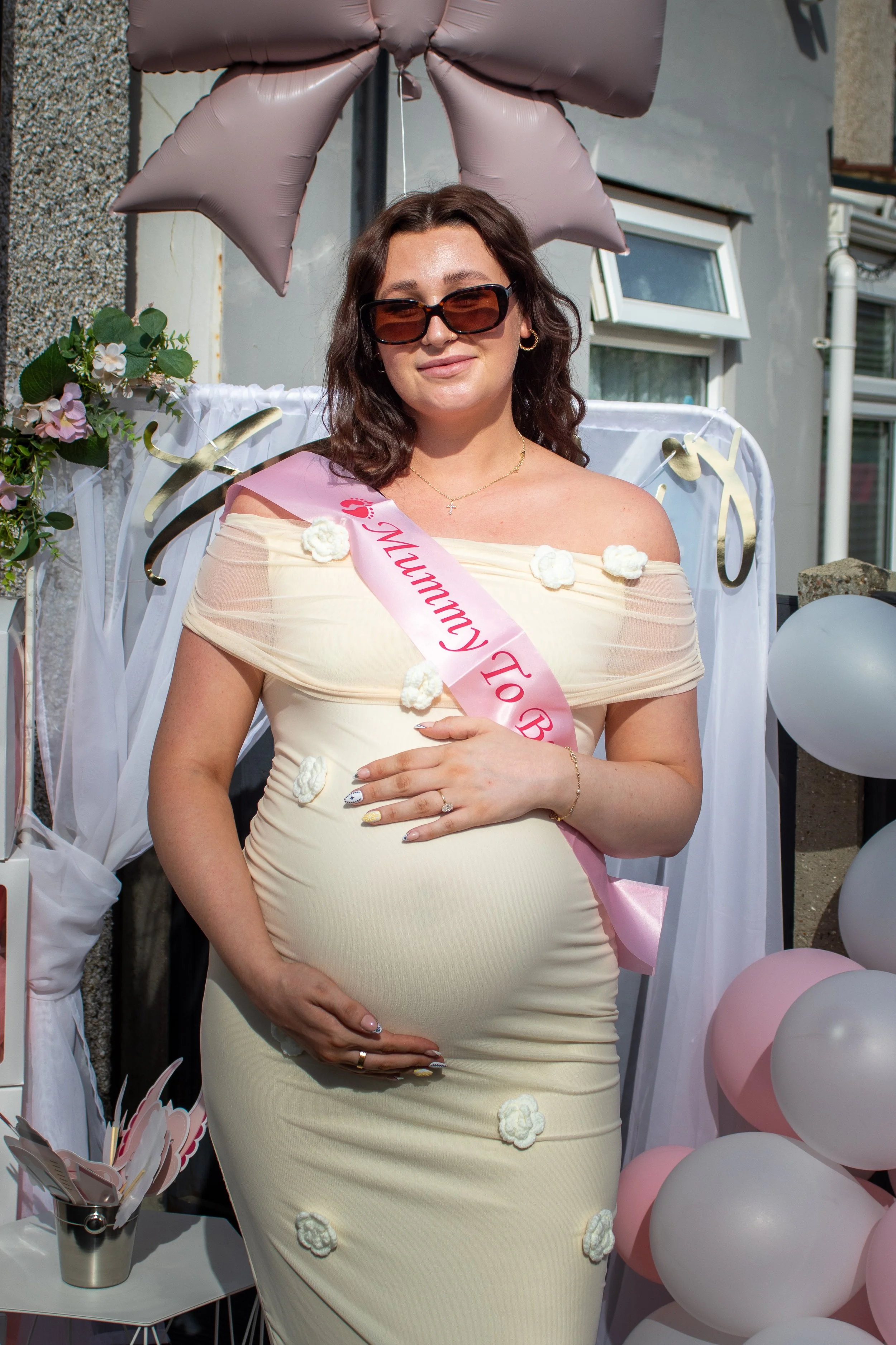 Pregnant woman wearing sunglasses and a off-shoulder cream dress decorated with white flowers, holding her belly, standing in front of a backdrop with pink and white balloons, flowers, and a large pink bow balloon.