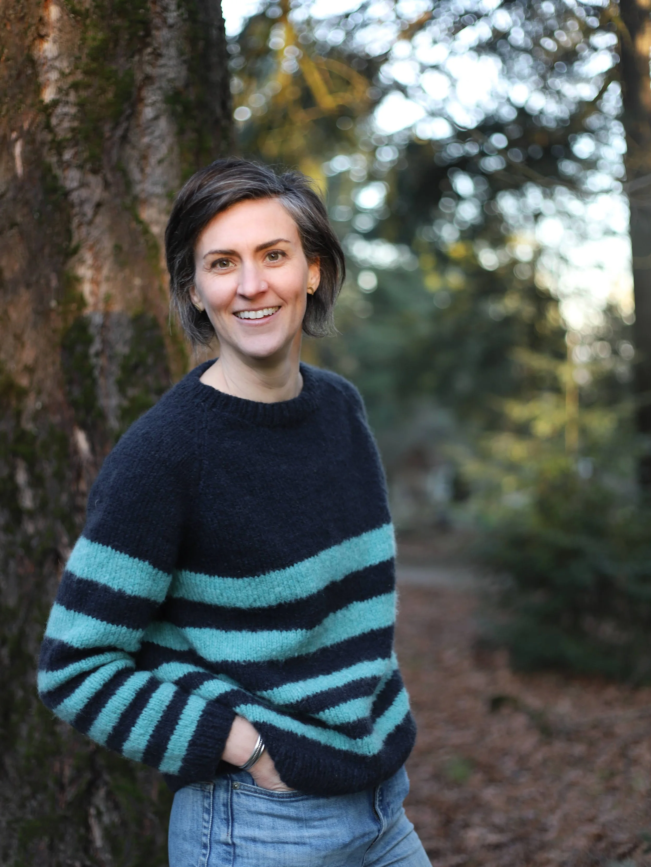A woman with short dark hair smiling outdoors, standing next to a tree with blurred trees and sky in the background, wearing a dark sweater with blue and teal horizontal stripes and jeans.