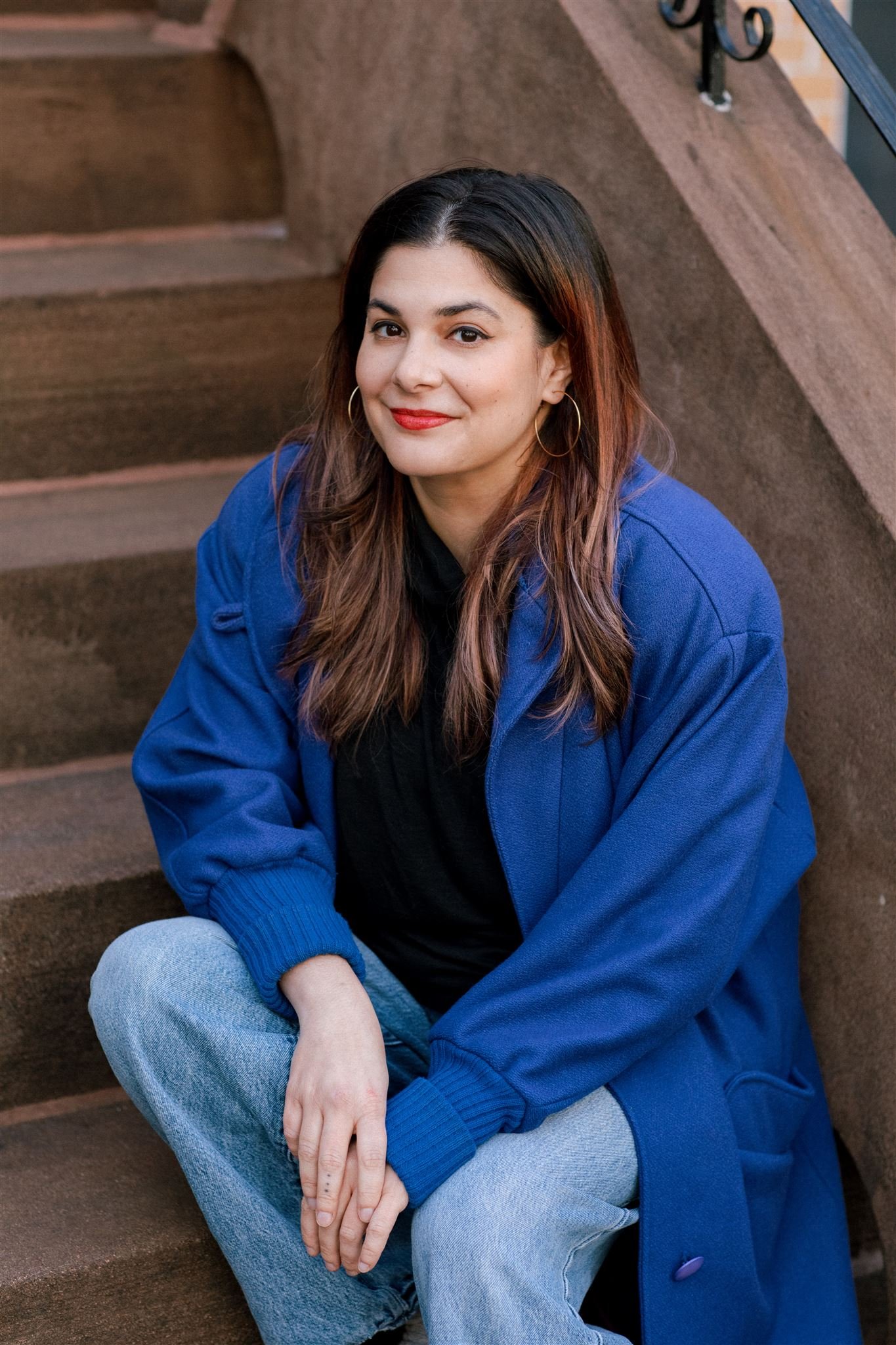 A young woman with brown hair, wearing a blue jacket and light gray jeans, sitting on outdoor stairs with a brown textured surface. She is smiling, has hoop earrings, and is looking at the camera.