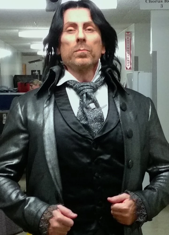 Acting studio instructor posing as Doctor Miracle in Tales of Hoffmann, wearing a black leather jacket, white shirt, and patterned tie, with long black hair..