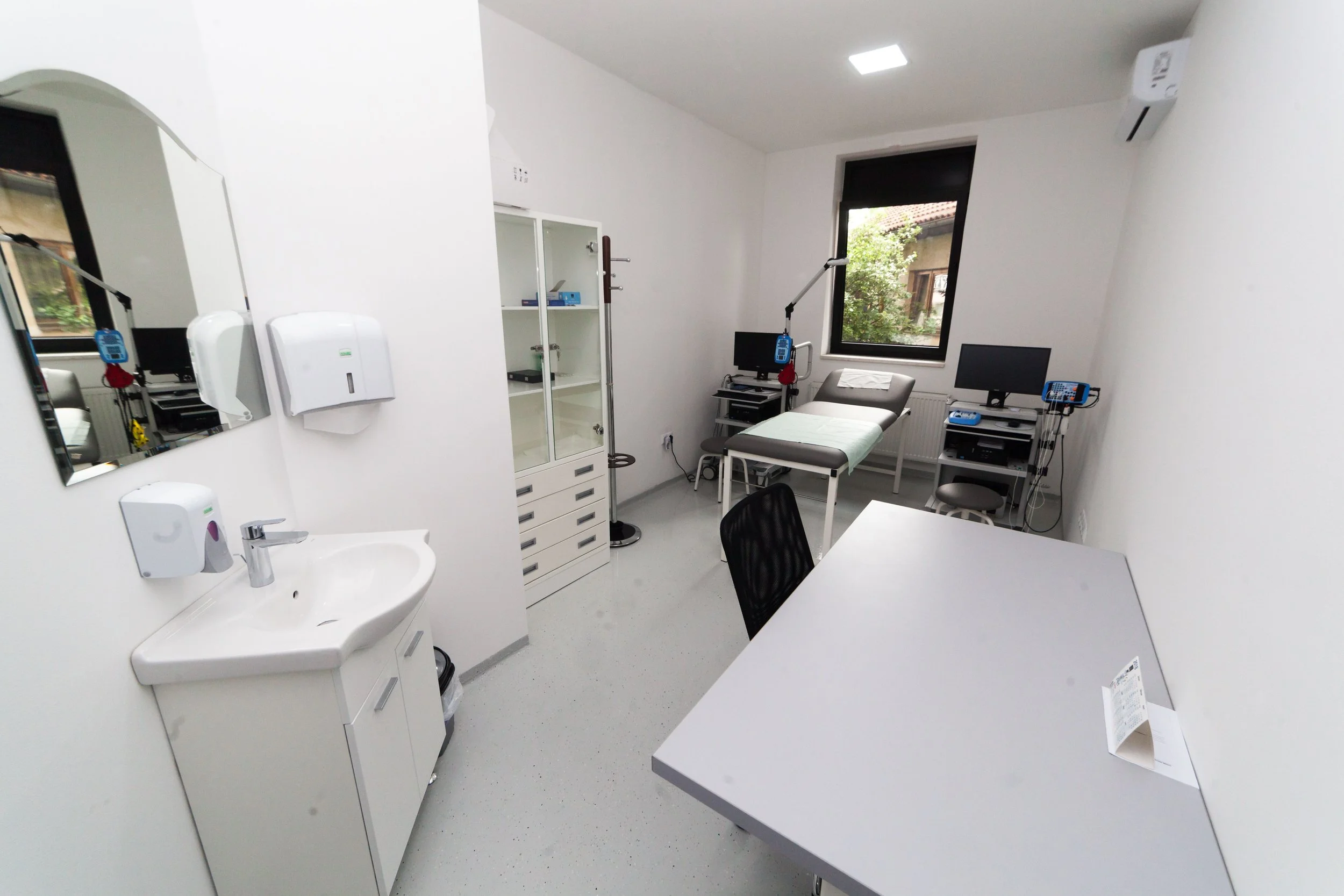A medical examination room with white walls, a window, examination bed, multiple computer monitors, medical equipment, and a sink.