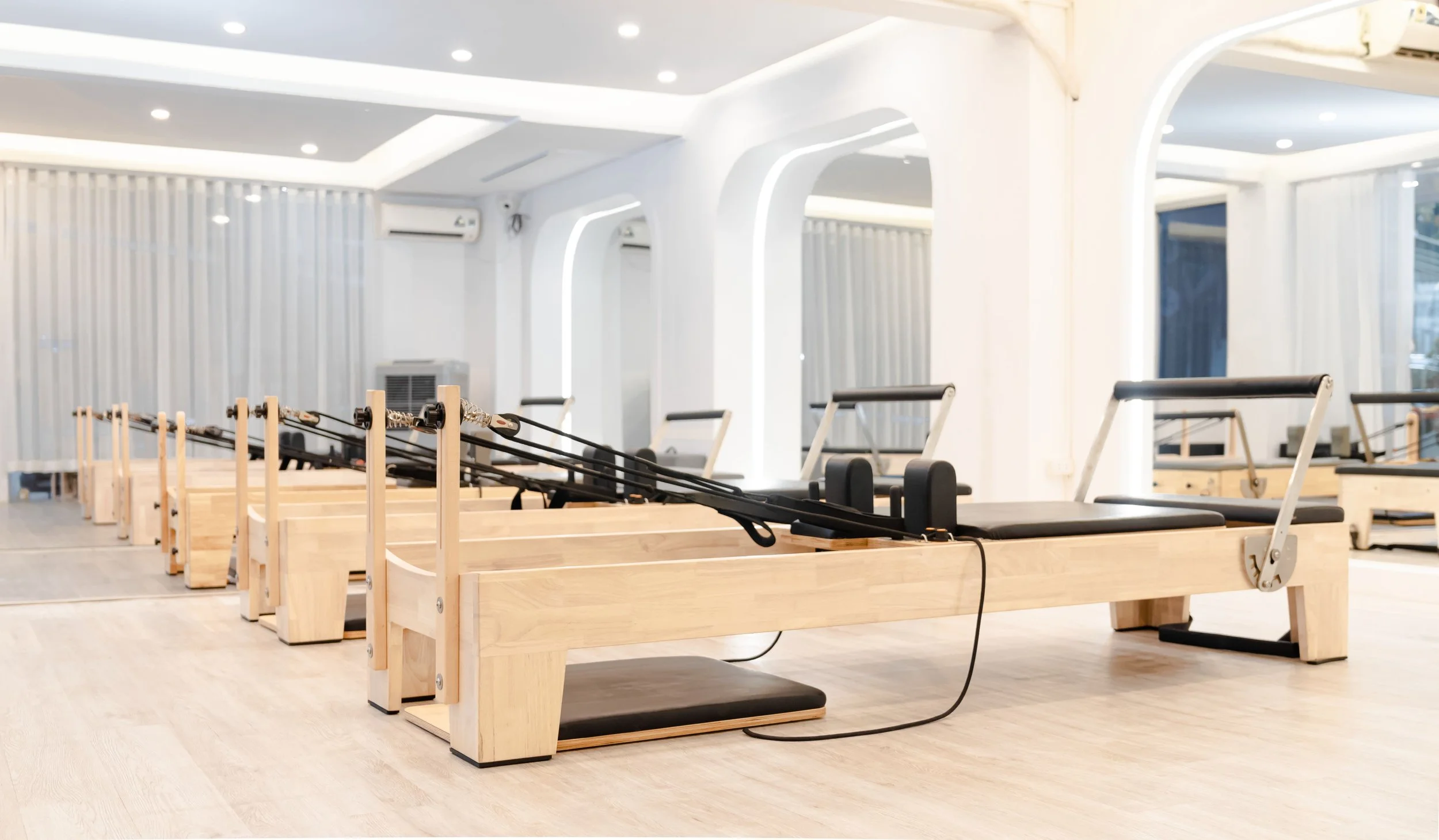 Pilates reformer machines in a bright, modern exercise studio with white walls, arched windows, and wood flooring.