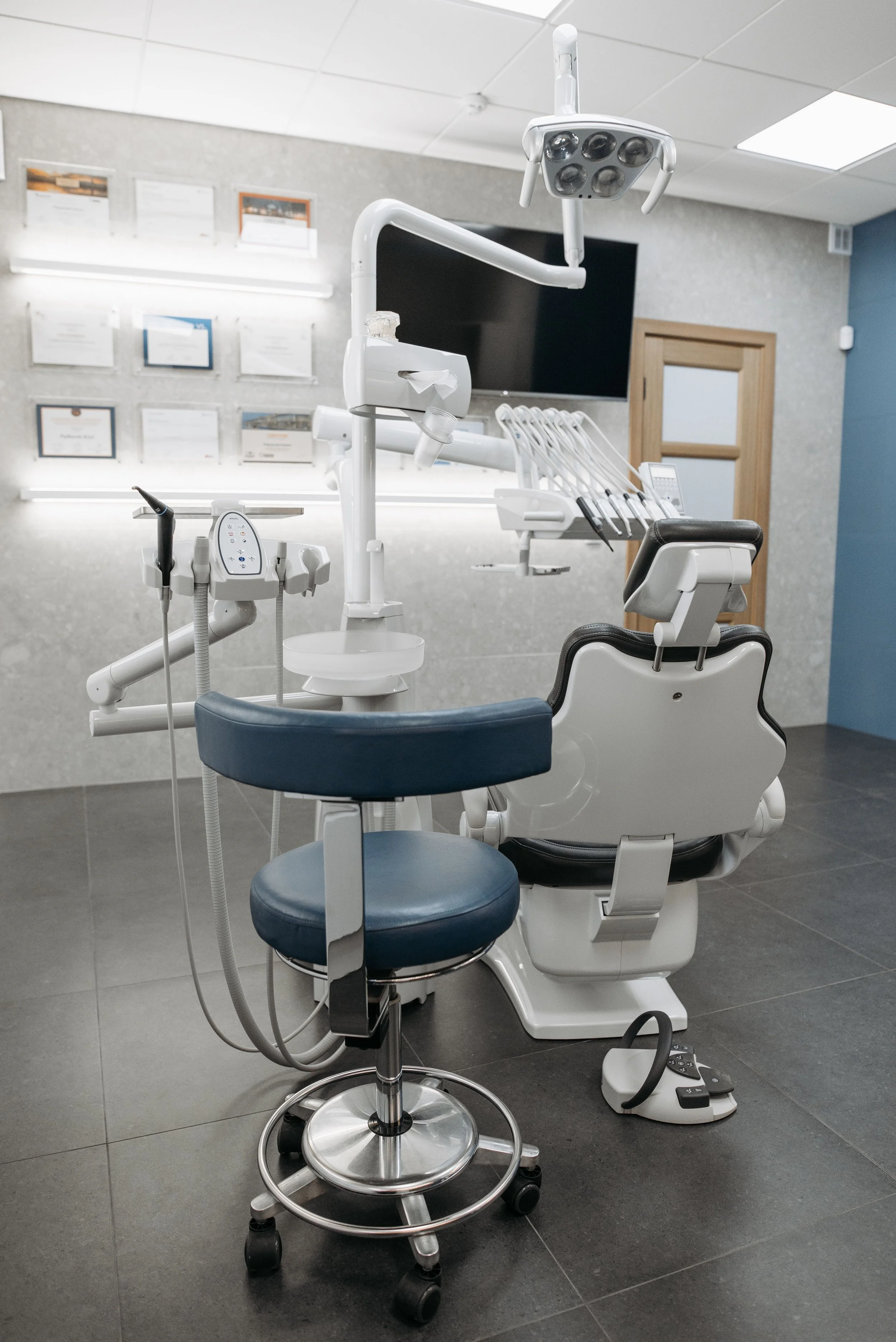 A modern dental or medical examination room with a dental chair, dental equipment, monitors, and wall certificates.