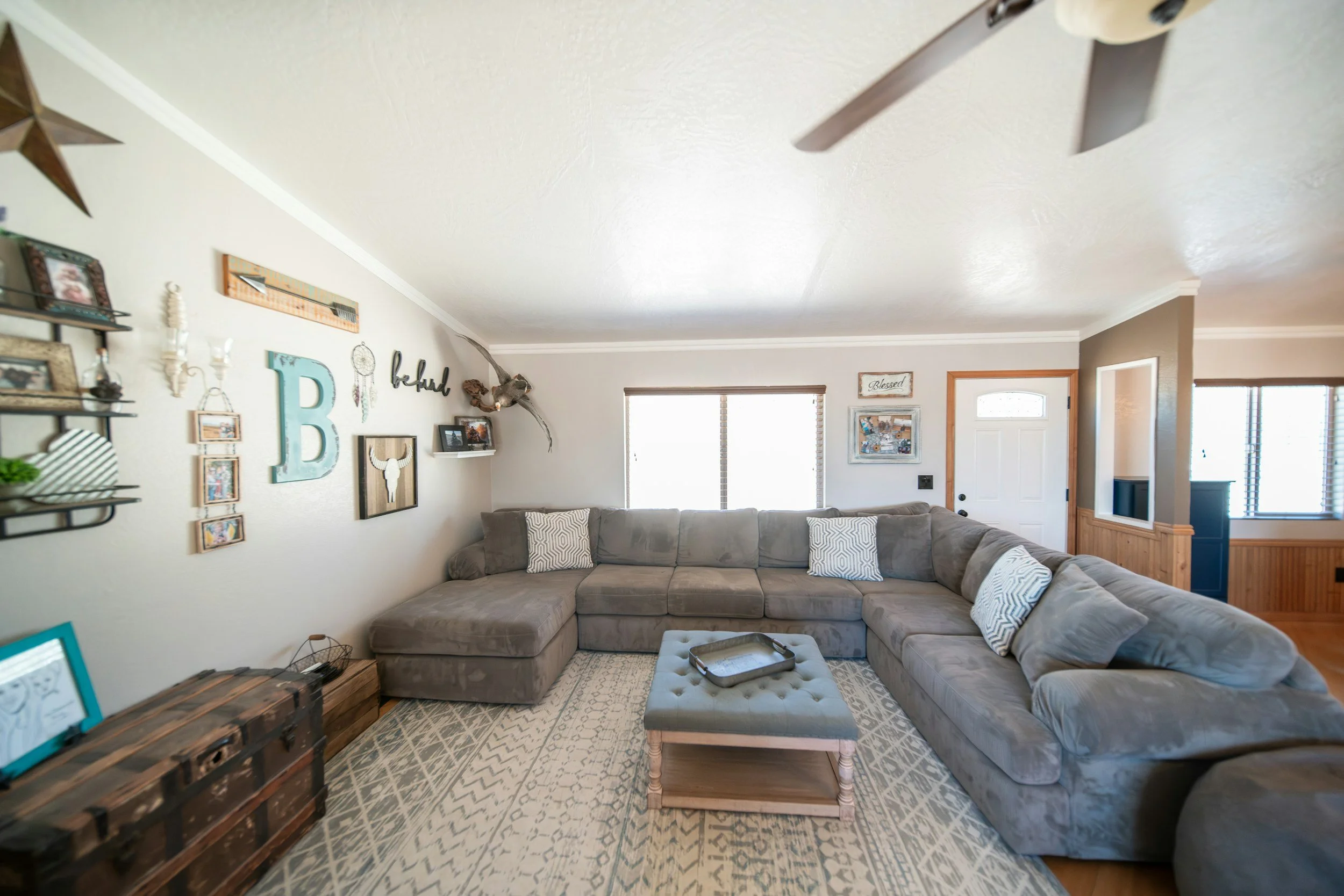 Living room with large gray sectional sofa, pillows, and a wooden coffee table, decorated with wall art, a window with blinds, and a front door.