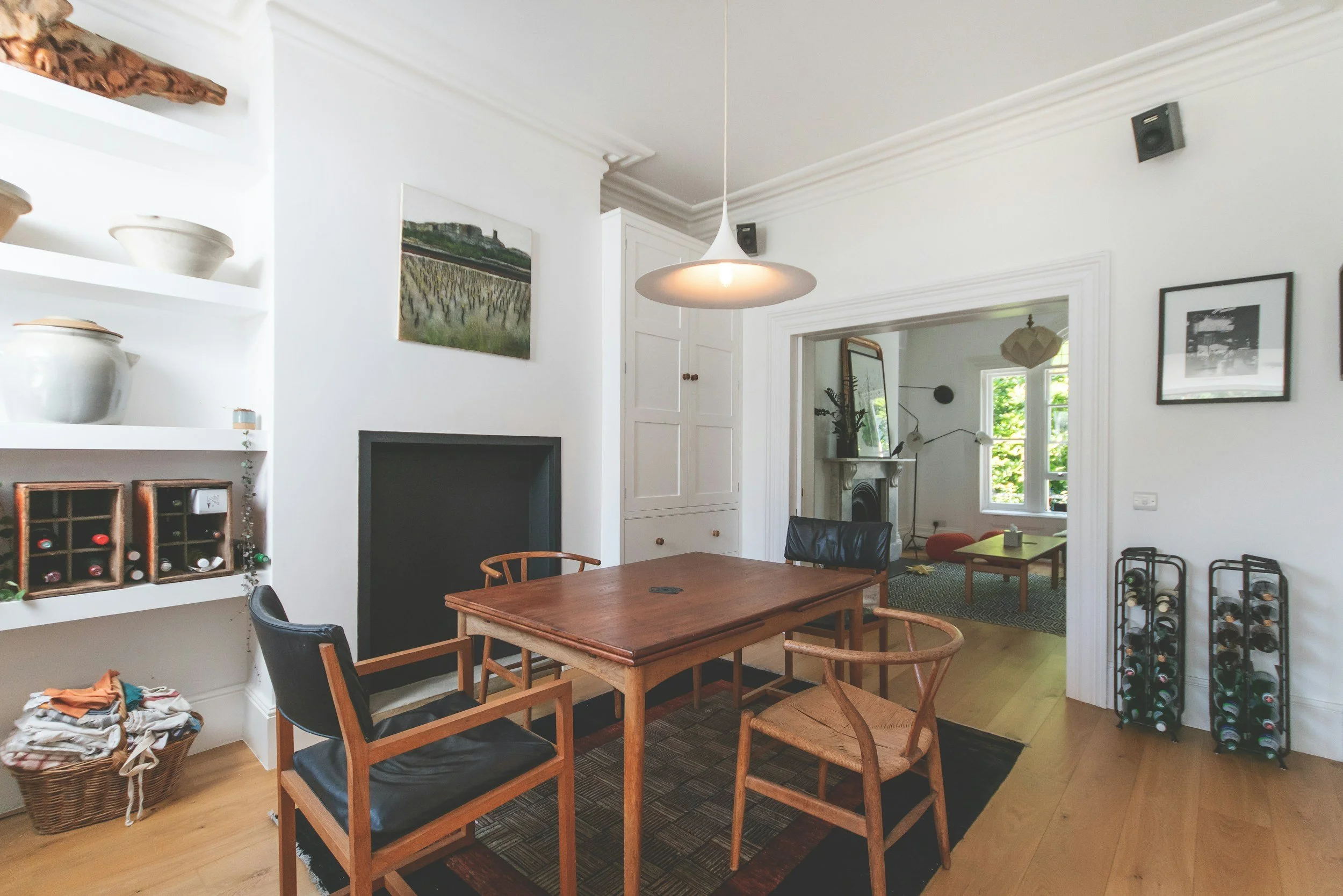 A dining room with a wooden dining table and six chairs, a wall with built-in white cabinets, framed artwork, a window, and a doorway leading to another room with a fireplace and a mirror. Shelves with pottery and wine bottles are also visible.