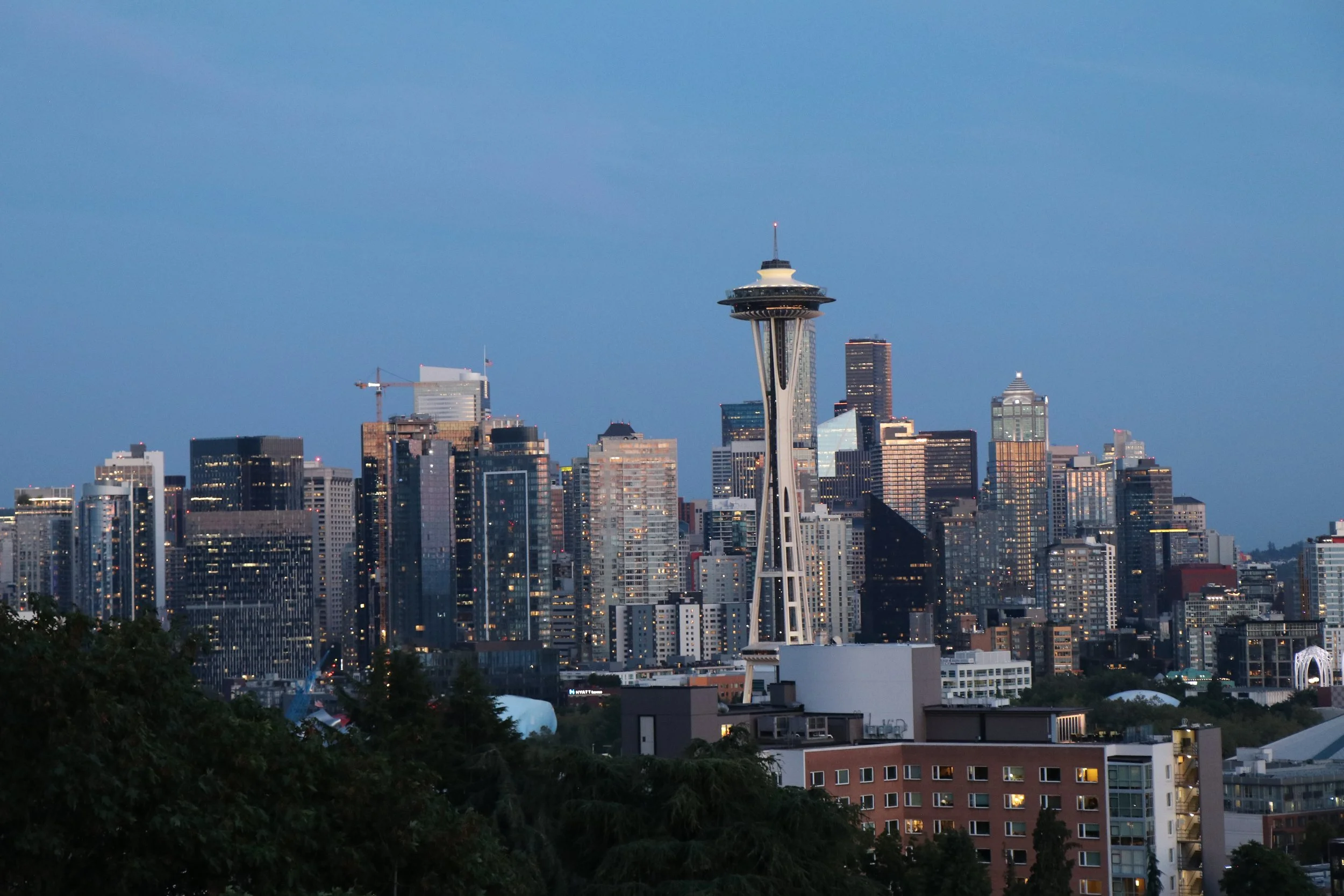 Dusk skyline of Seattle, Washington, with the Space Needle in the center, surrounded by tall buildings and a few construction cranes, under a mostly clear sky.