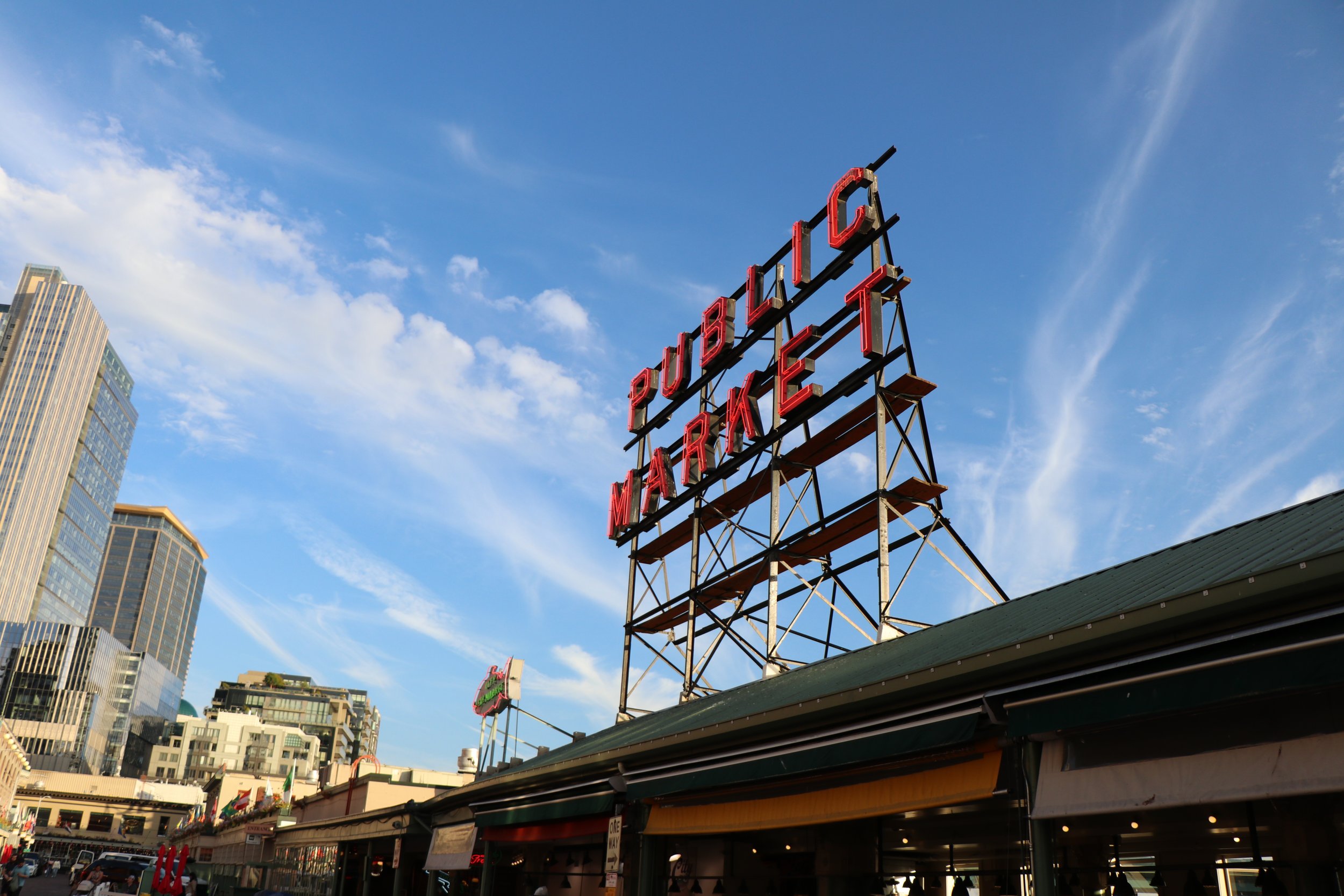 View of a city street with a neon sign that reads 'Public Market' against a blue sky with some clouds. Tall buildings are visible in the background.