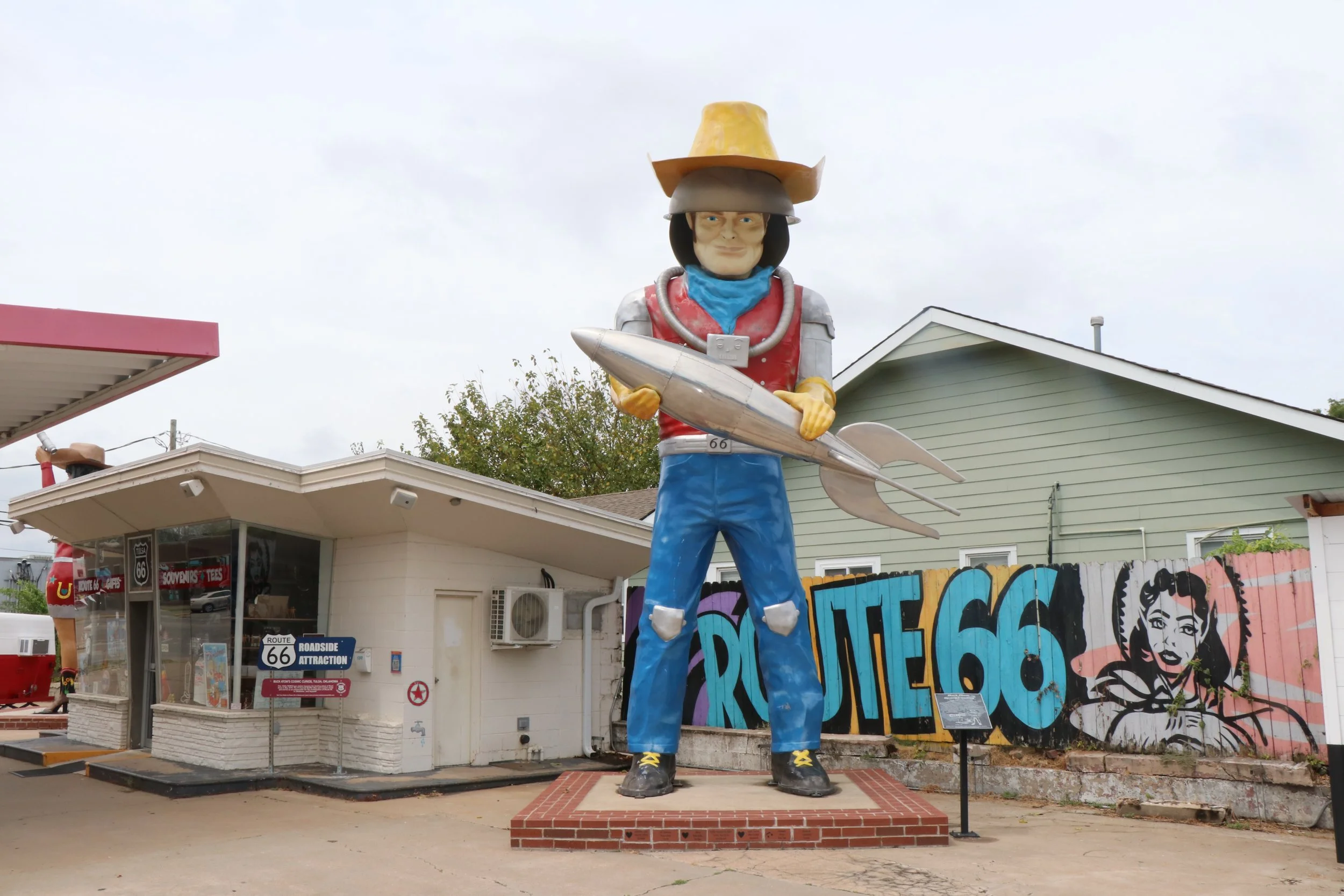 Large colorful statue of a person wearing a cowboy hat, holding a surfboard, and standing in front of a building with graffiti art and Route 66 signs.