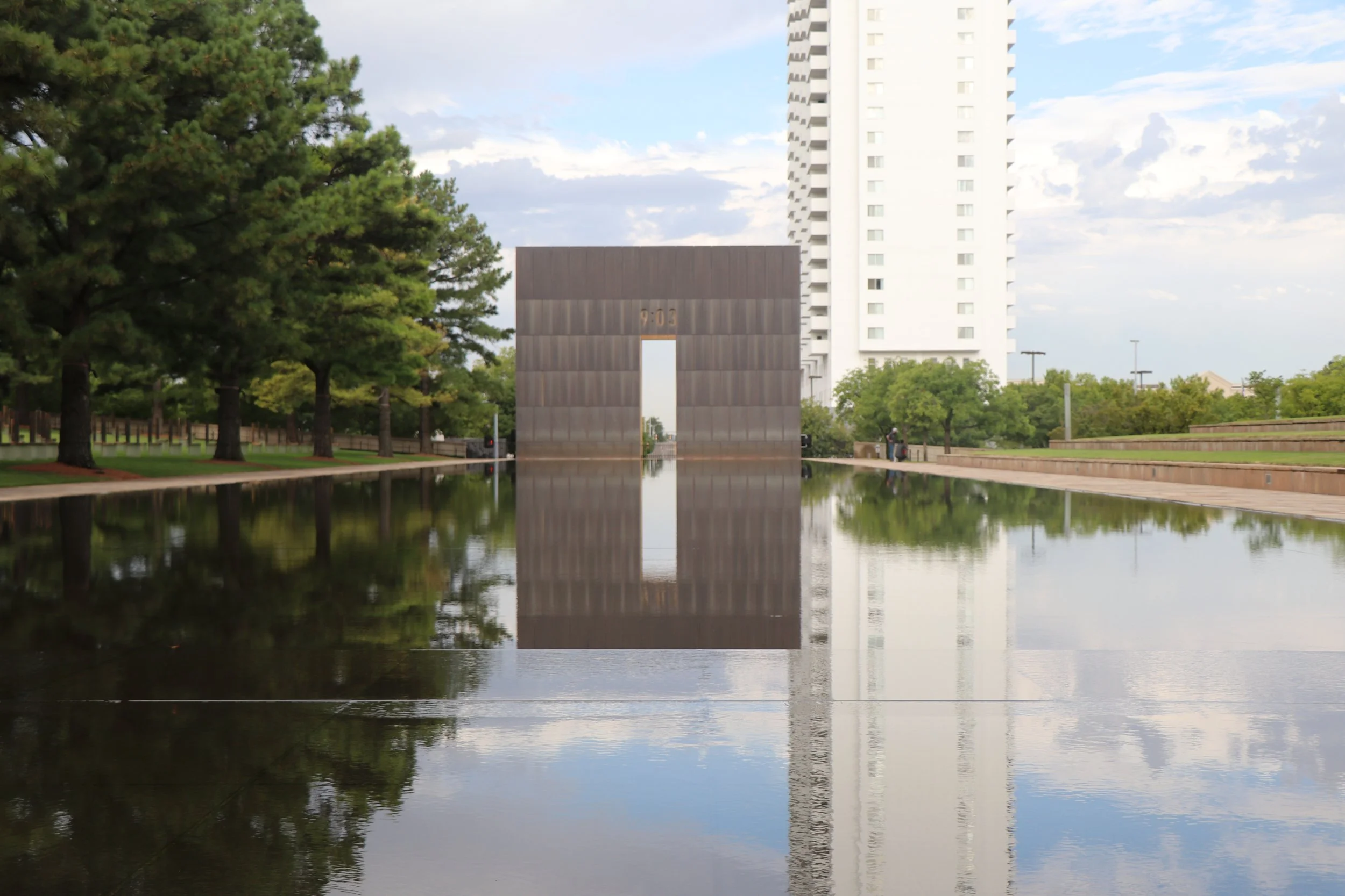 A park with green trees lining a reflective water feature, with a modern high-rise building and a large welcome gate reading '9-03' in the background under partly cloudy sky.