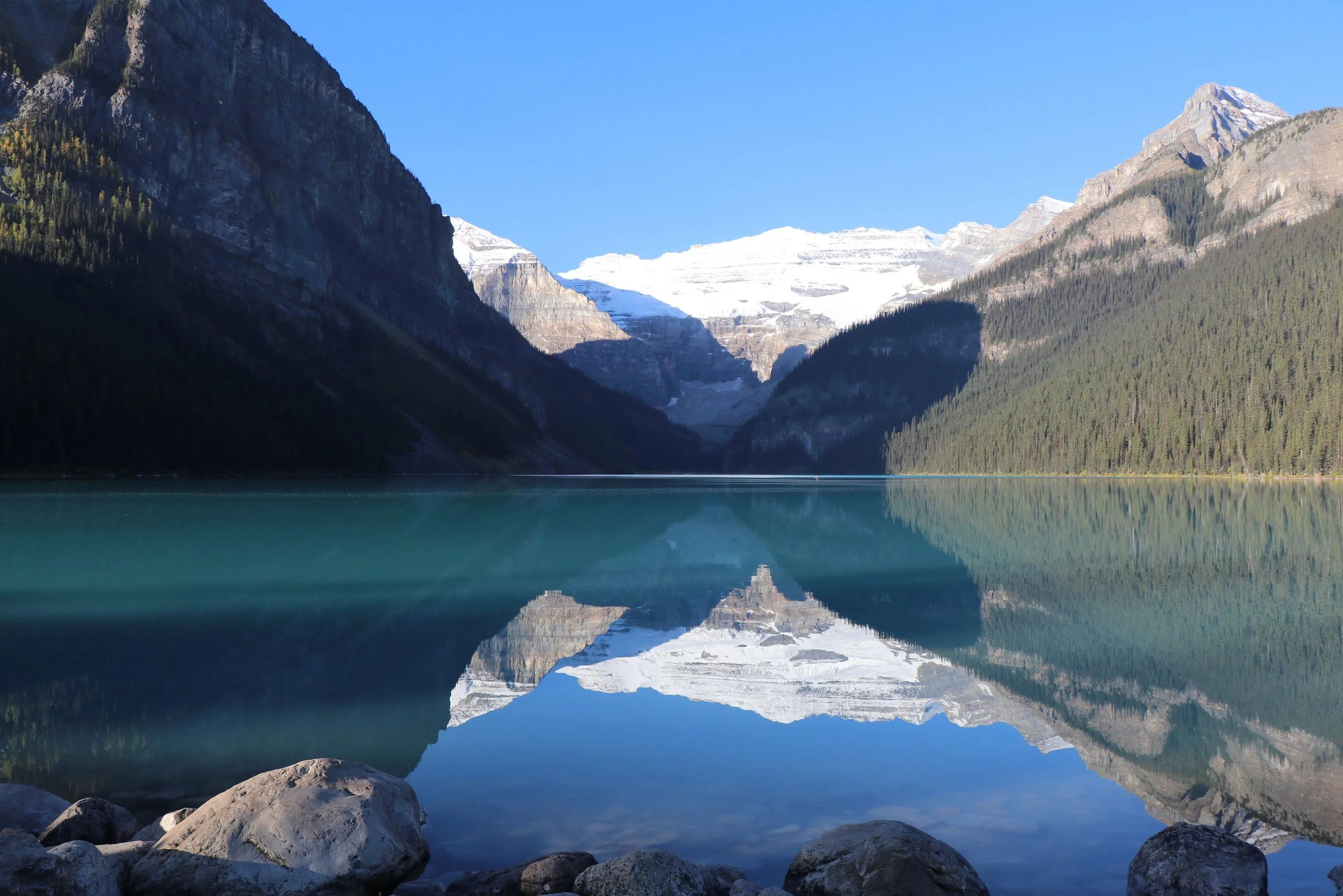 A serene mountain lake reflects snow-capped peaks and forested mountains under a clear blue sky, with rocks along the shoreline.