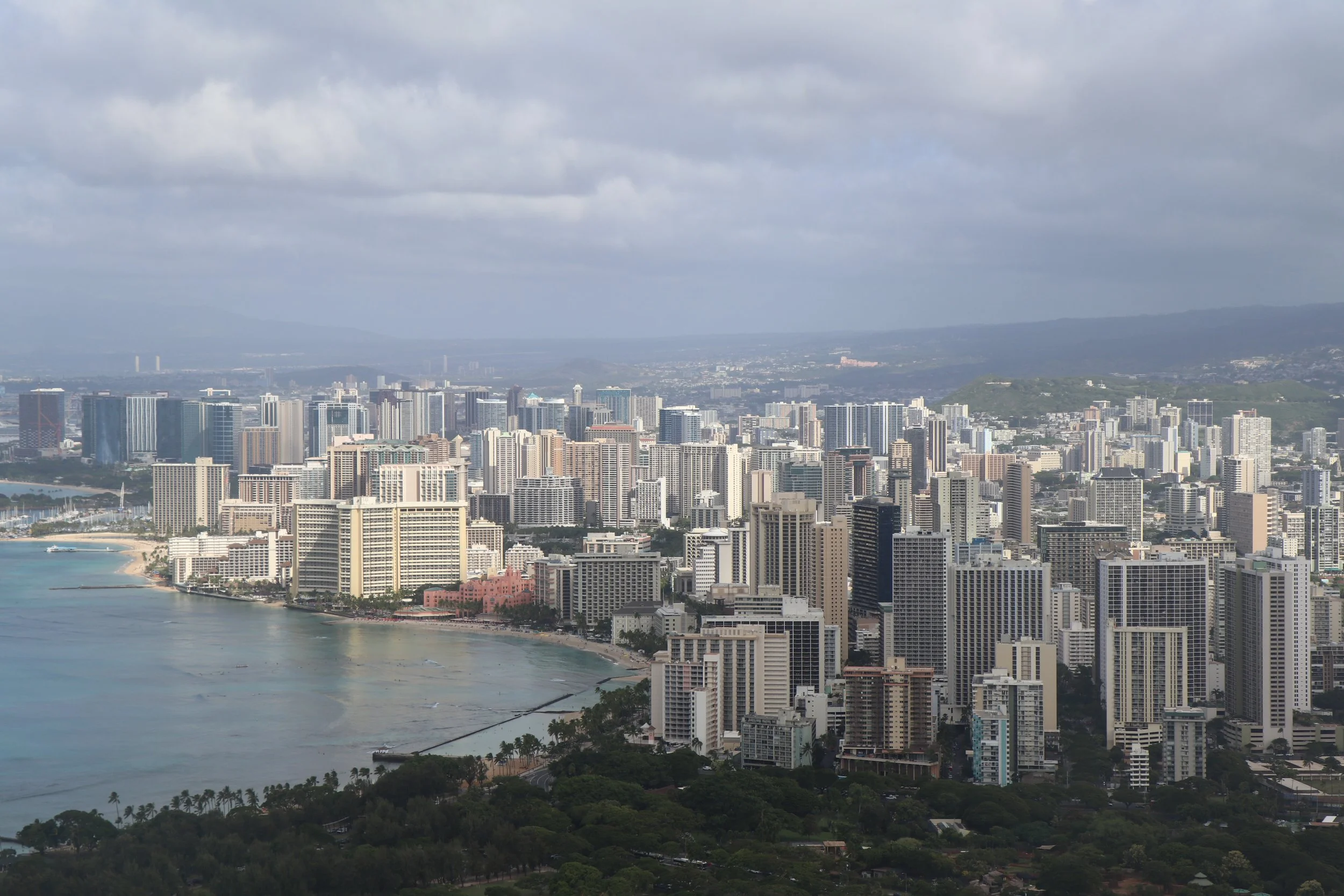 A city skyline with numerous high-rise buildings along the waterfront, with a beach visible in the foreground and mountains in the background under a cloudy sky.