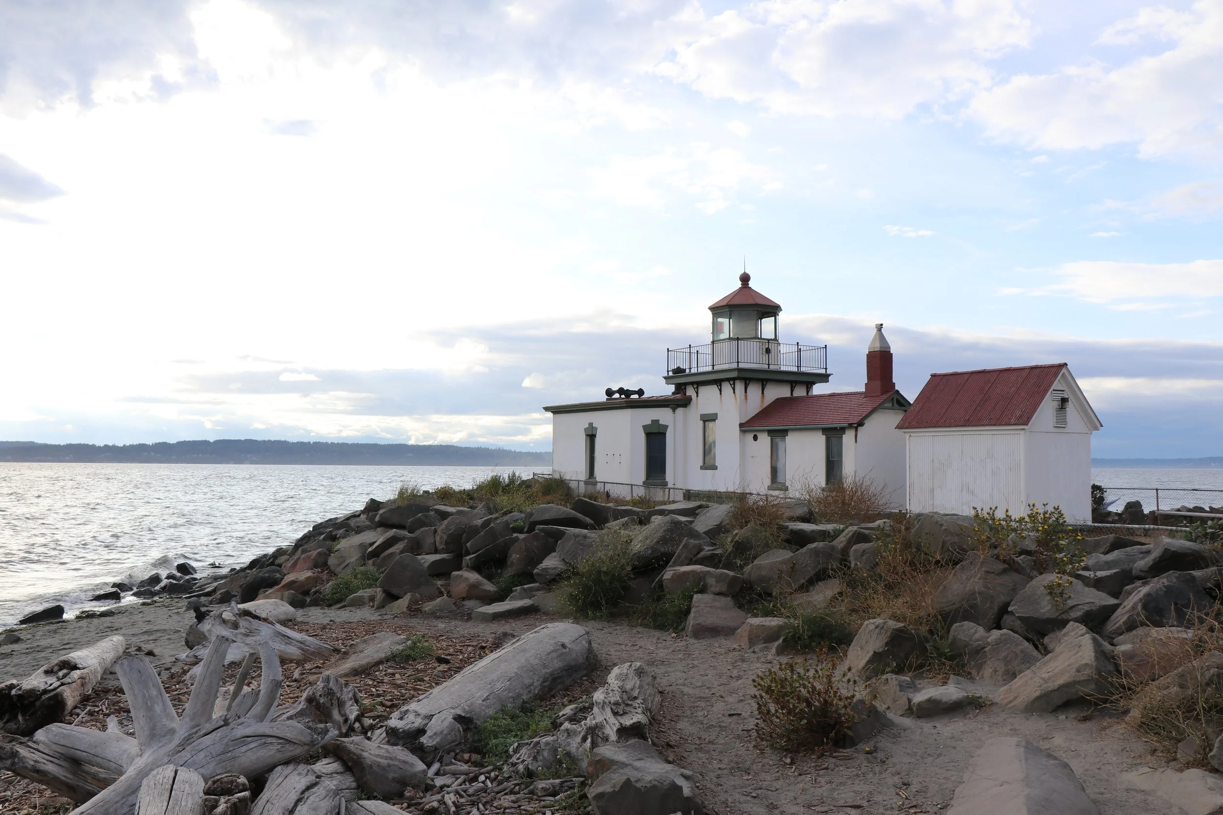 A lighthouse with a small attached building is situated on rocky shore by the water under a cloudy sky.