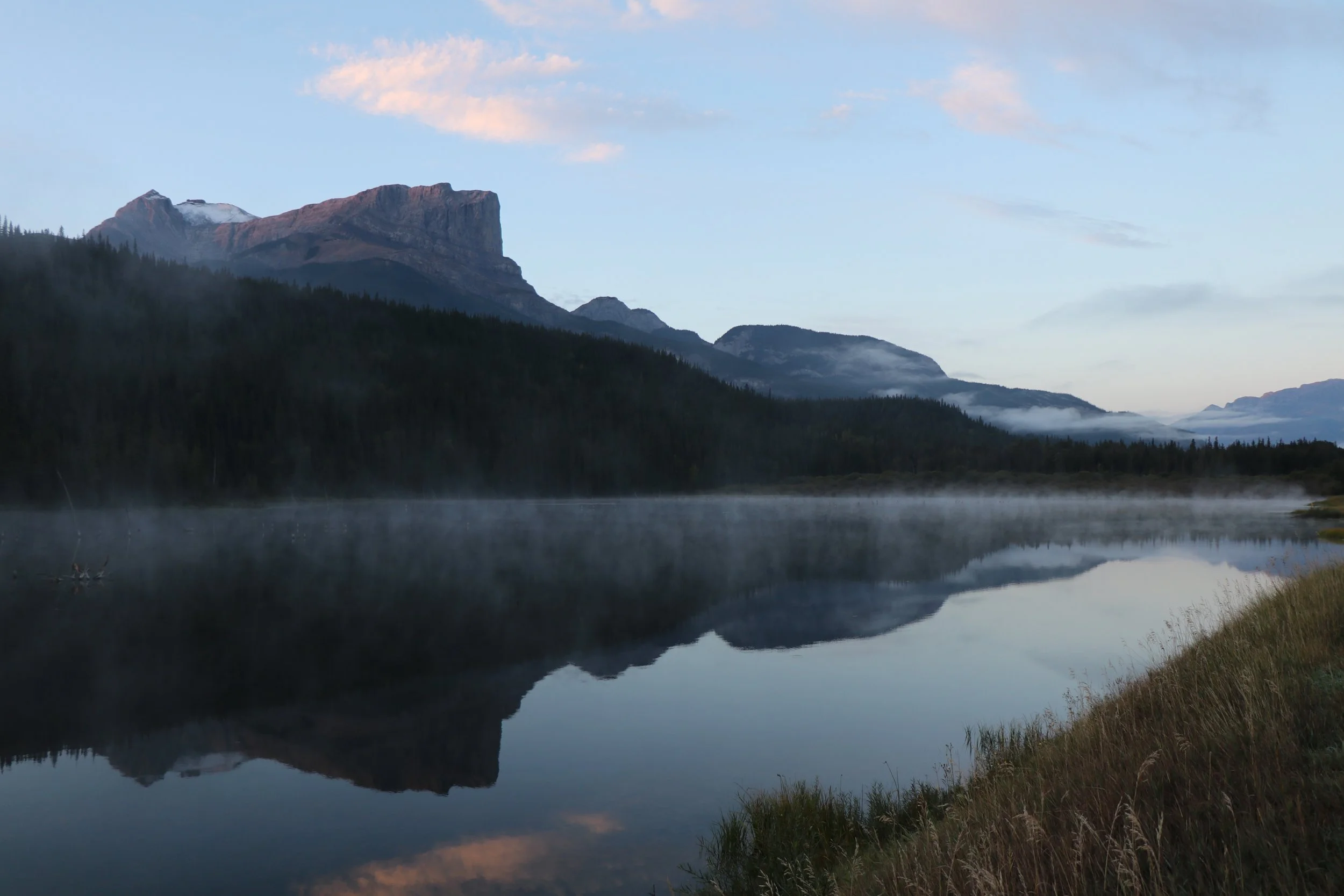 A tranquil mountain landscape with a calm lake reflecting the mountains and sky, featuring a large rocky mountain with some snow, a forested hillside, and a partly cloudy sky at dawn or dusk.