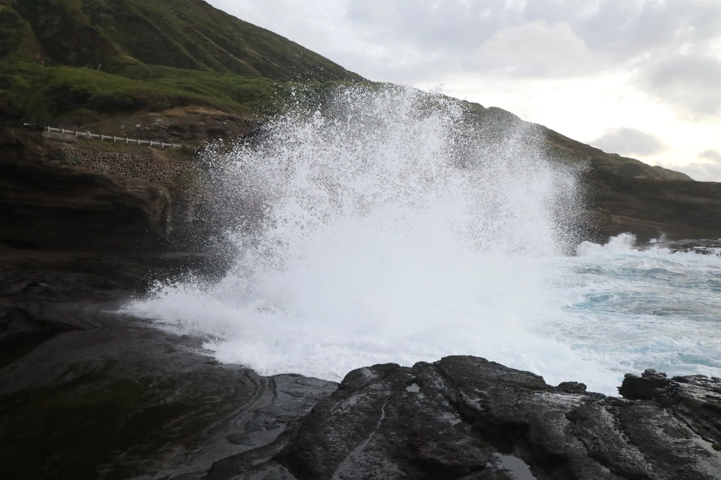 Waves crashing against rocky shoreline with green hills and cloudy sky in the background.