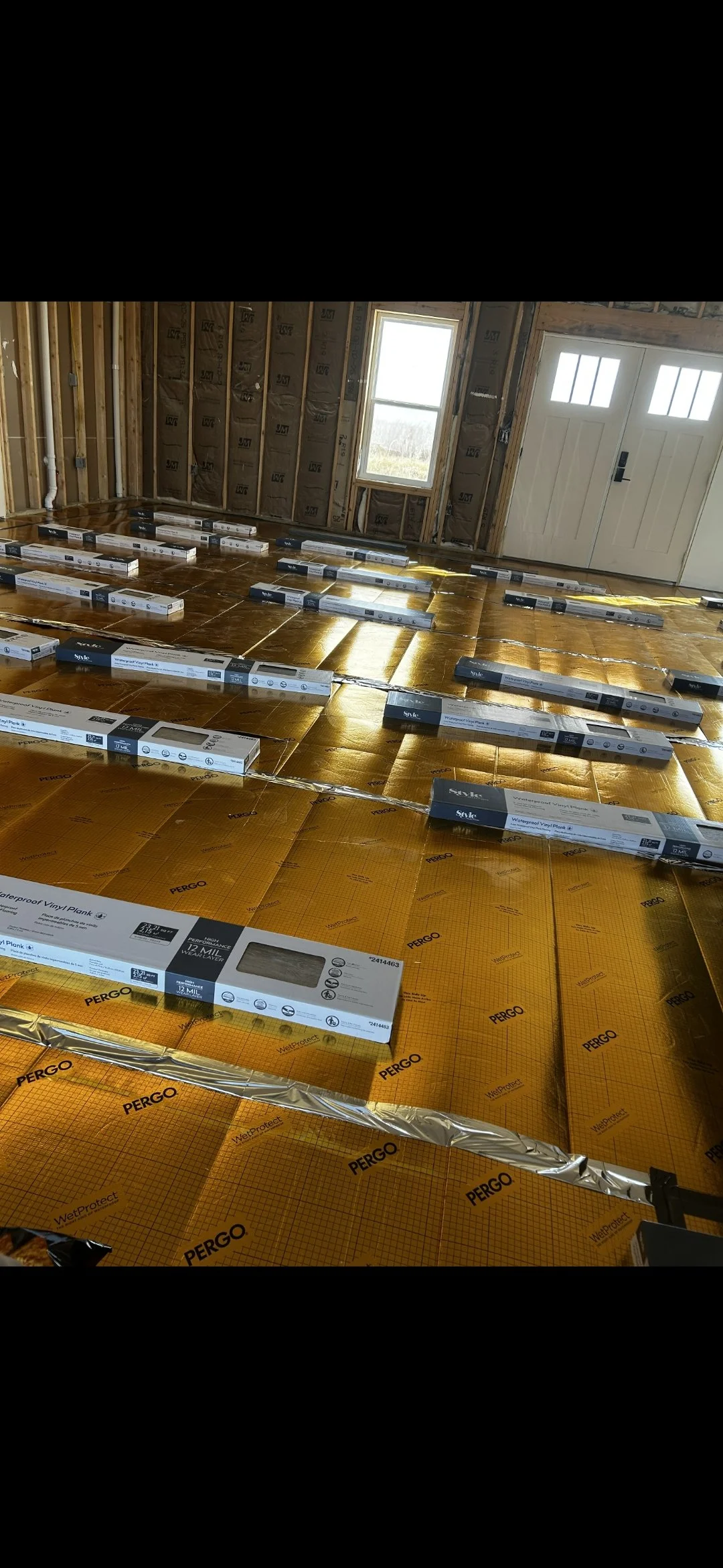 A room under construction with yellow waterproof vinyl planks on the floor, organized in rows, and partially installed. The walls are unfinished with exposed studs, insulation, and a window and a garage door visible.