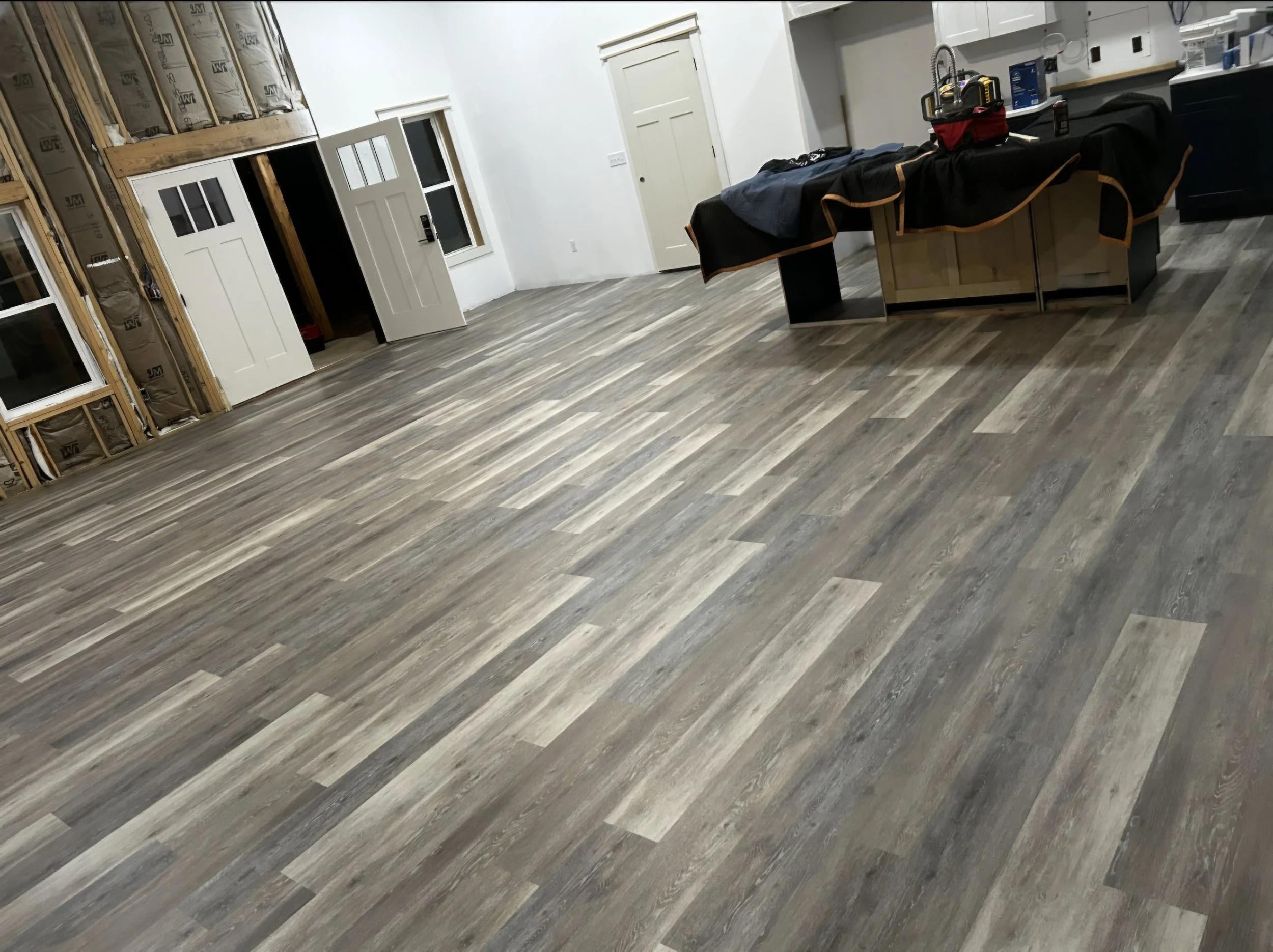 An unfinished room with exposed insulation, white walls, and wooden flooring. A door is open, revealing storage space with construction supplies. There is a countertop with a red toolbox, a black cloth covering part of it, and various small items. Th