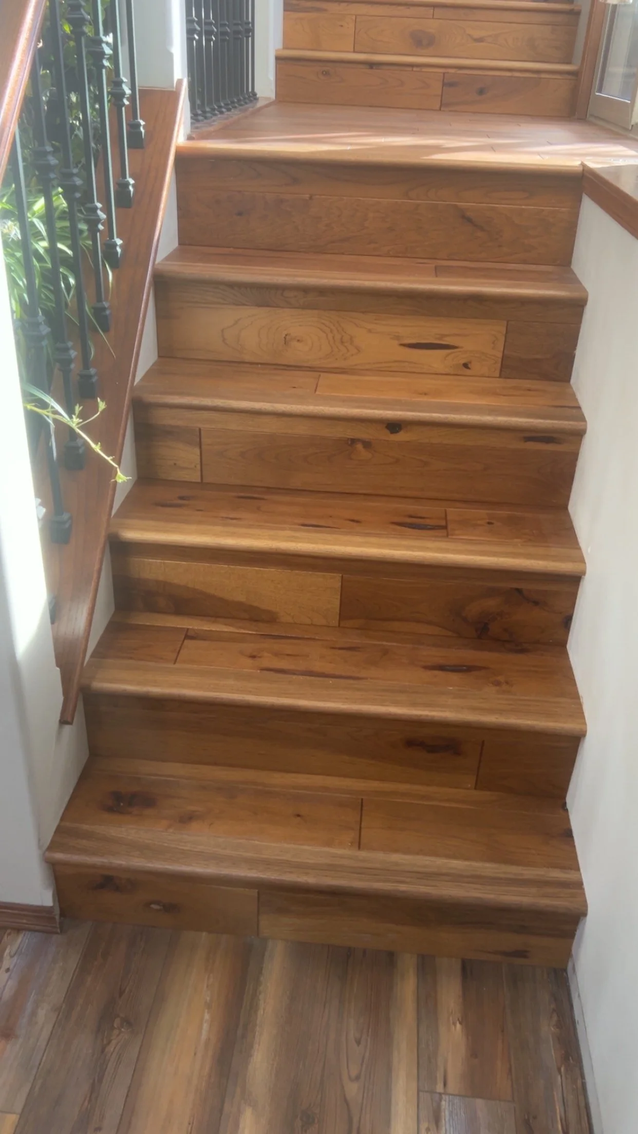 Wooden staircase with seven steps and a wooden handrail on the left side, with a black metal railing next to it. The stairs are in a well-lit area with a view of greenery outside.