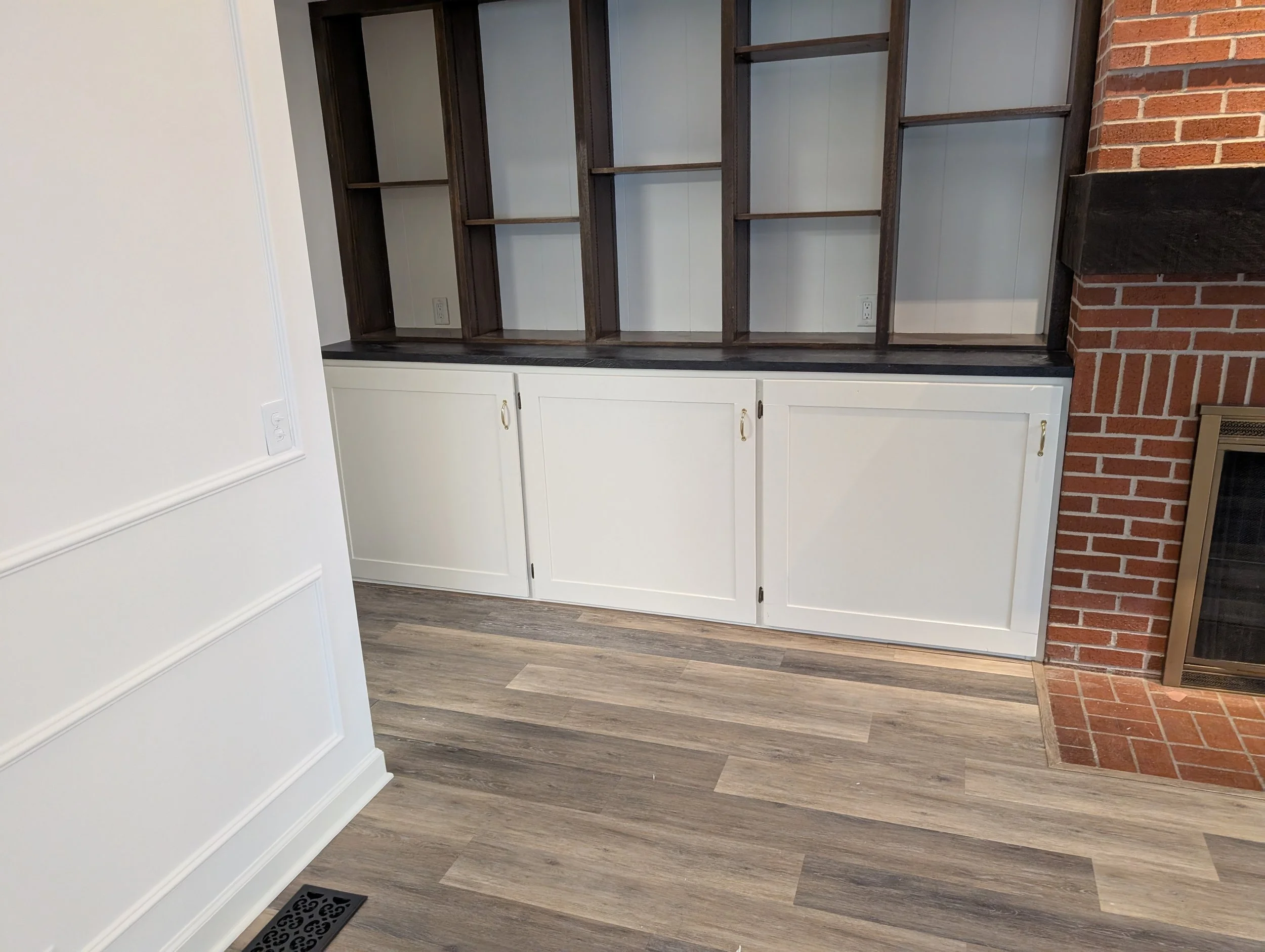 Interior of a room with a wooden built-in shelving unit, white cabinets below, a brick wall to the right, and a floor vent near a white wall with paneling.