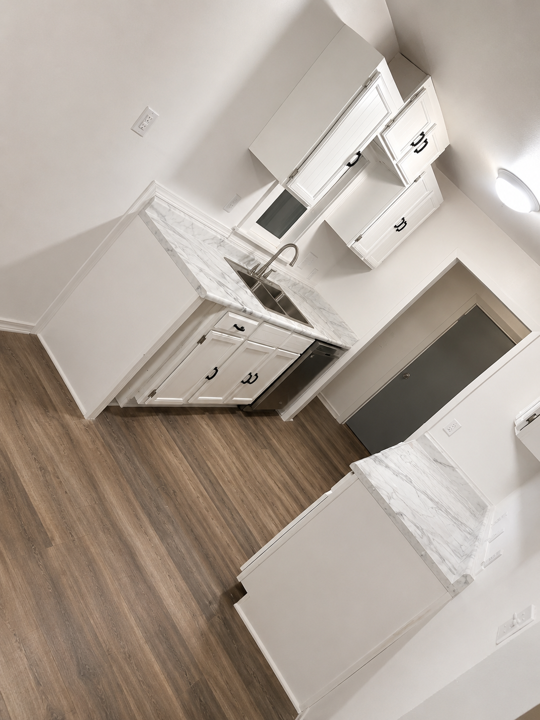 Empty white kitchen with marble countertops, white cabinets with black handles, stainless steel sink, hardwood floor, gray door, and white walls.