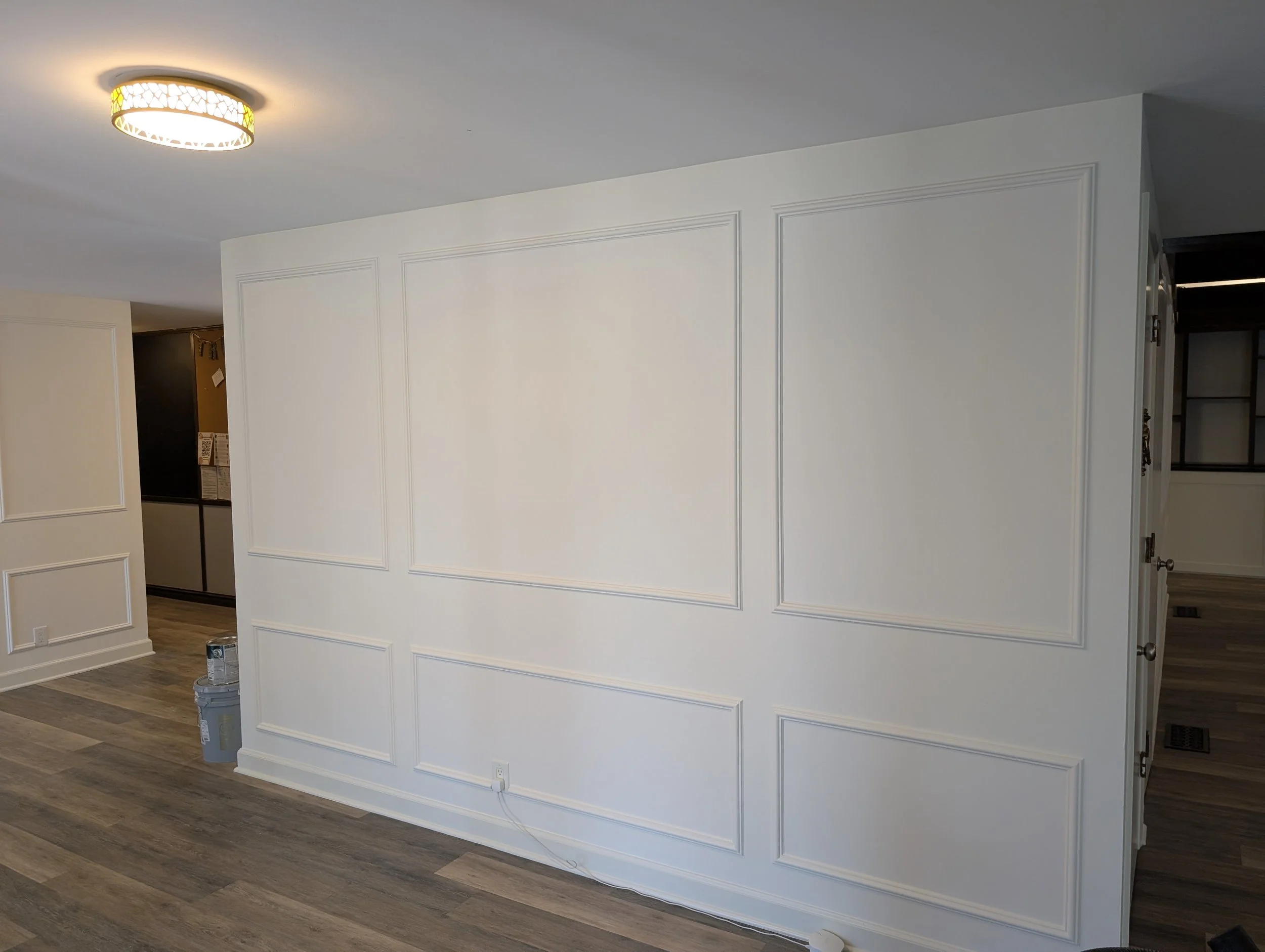 Interior of a room with white walls, decorative wall molding, and dark wood flooring. Part of a kitchen area is visible to the right, and paint cans are on the floor near the wall.