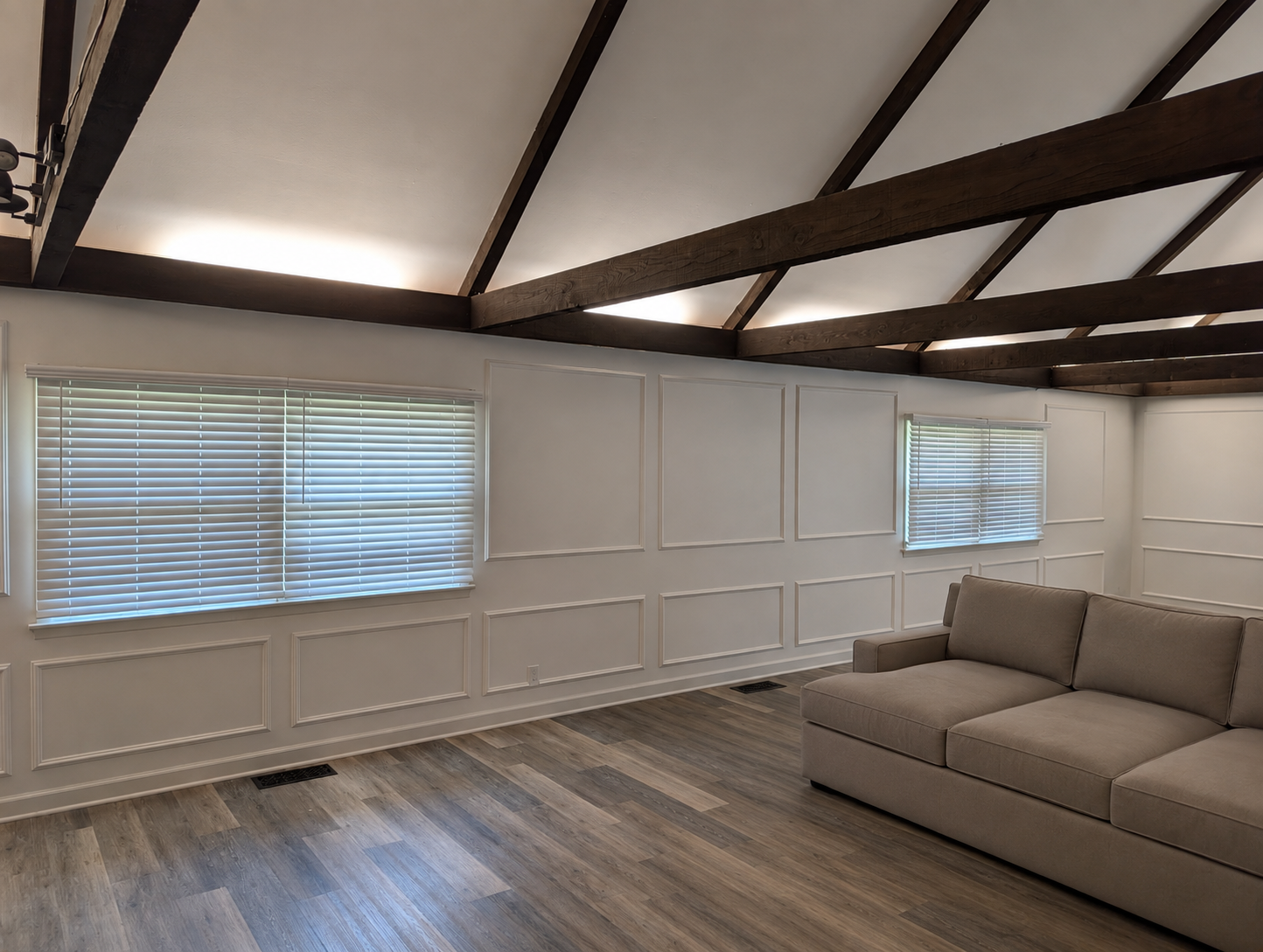 A living room with light hardwood flooring, white paneled walls, two windows with white blinds, and a beige sofa. The ceiling has exposed dark wooden beams and is vaulted with soft lighting.
