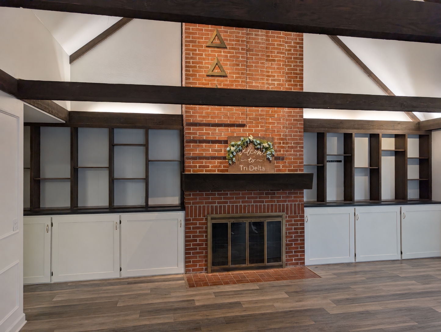 Living room with brick fireplace, dark wood beams, bookshelves with open and closed cabinets, wood flooring, and a welcome sign with floral decoration on the mantle
