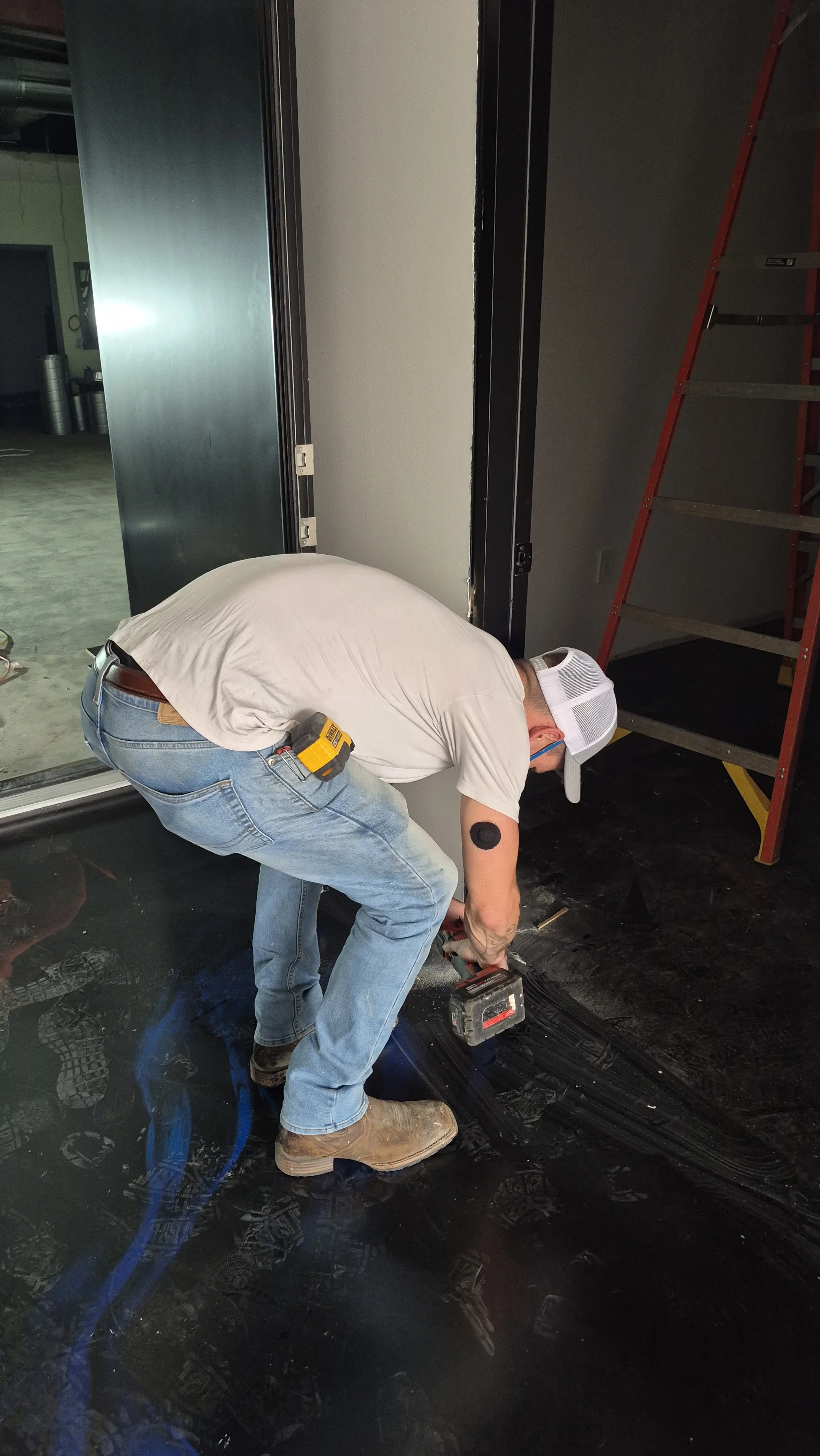 A man wearing a white shirt, blue jeans, and a white cap is working on the floor with tools, wearing sunglasses and a tool belt, in an industrial or warehouse setting with a ladder and open space in the background.