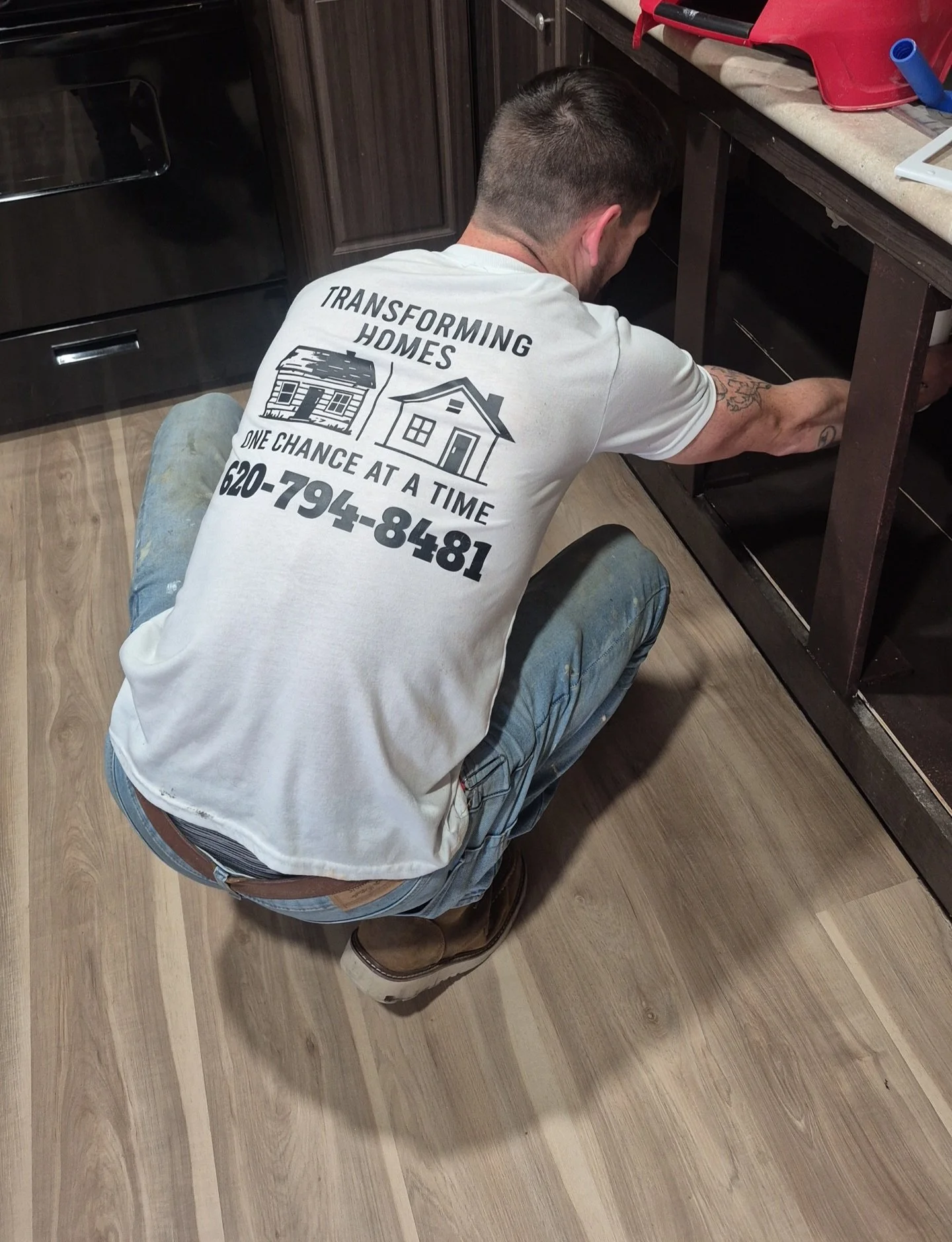 A man with short dark hair and tattoos kneeling in a kitchen, working under a dark wooden cabinet near a countertop. He is wearing a white T-shirt with black text and images promoting home transformation services.