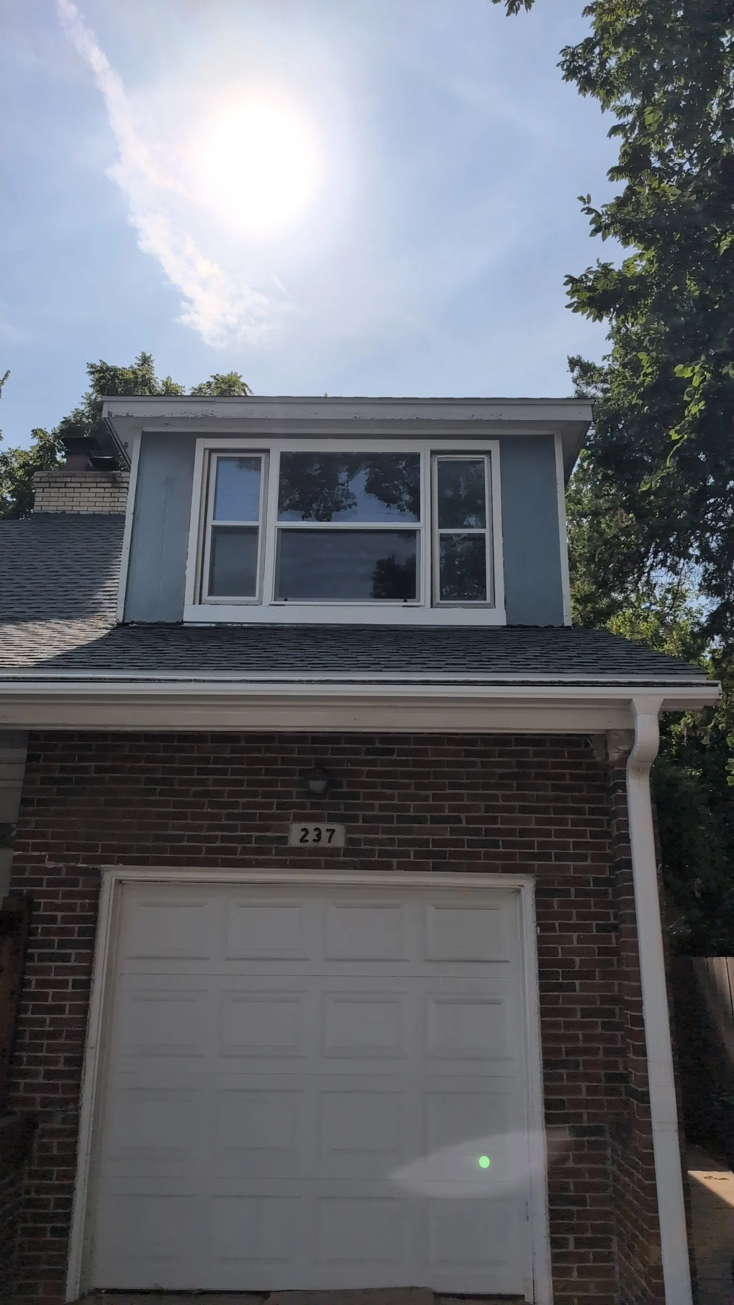 The image shows the front view of a two-story house with a brick facade, a white garage door, and a large window on the upper floor. The house number 237 is visible above the garage door. The roof is shingled, and there are trees on both sides of the house. The sky is mostly clear with the sun shining brightly.