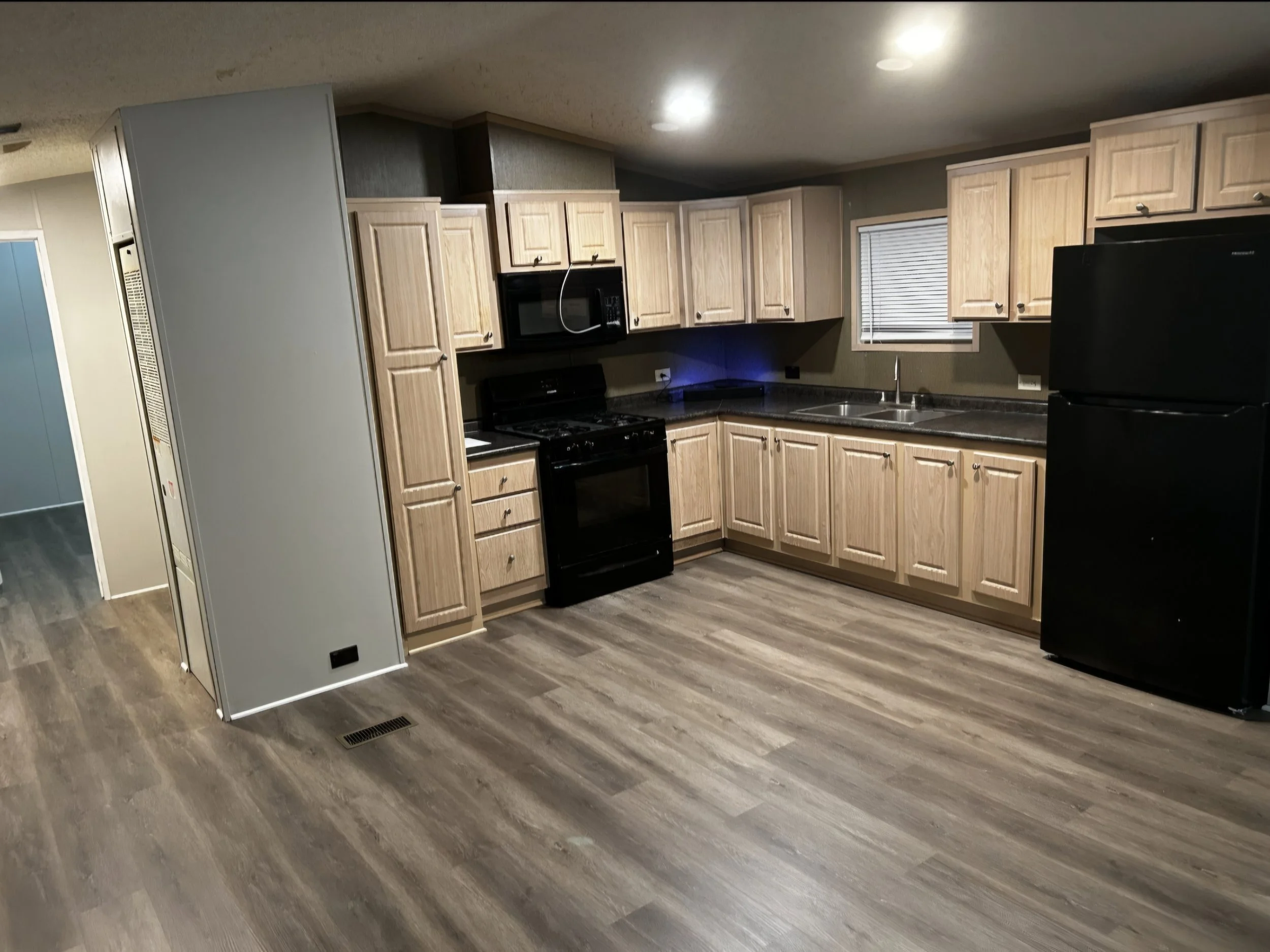 Kitchen with light wooden cabinets, black appliances, dark granite countertops, a small window with blinds, and a black refrigerator. The flooring is a dark wood or laminate.